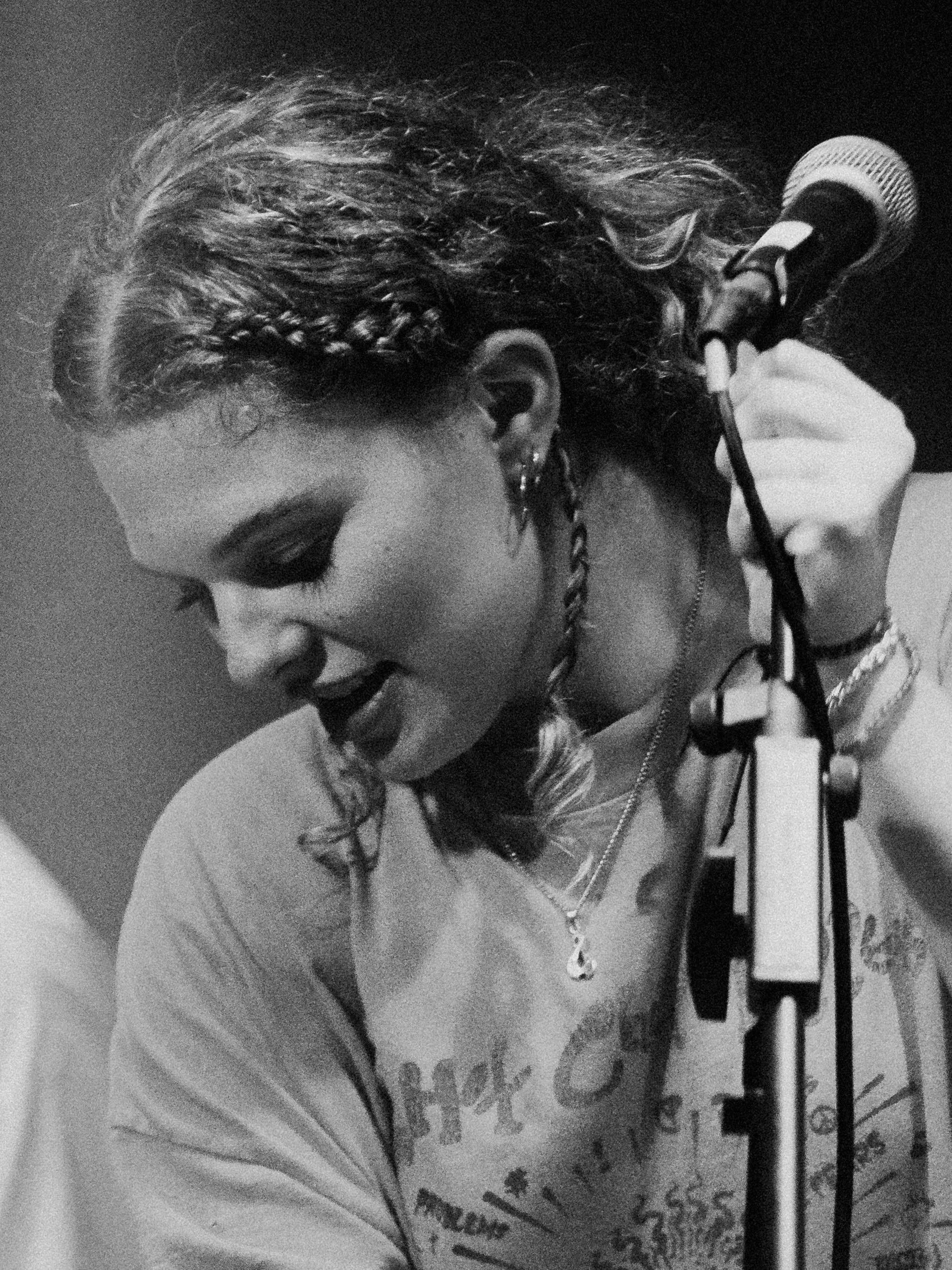 A woman singing into a microphone while holding a music stand, with curly hair, dark lipstick, earrings, and layered necklaces.