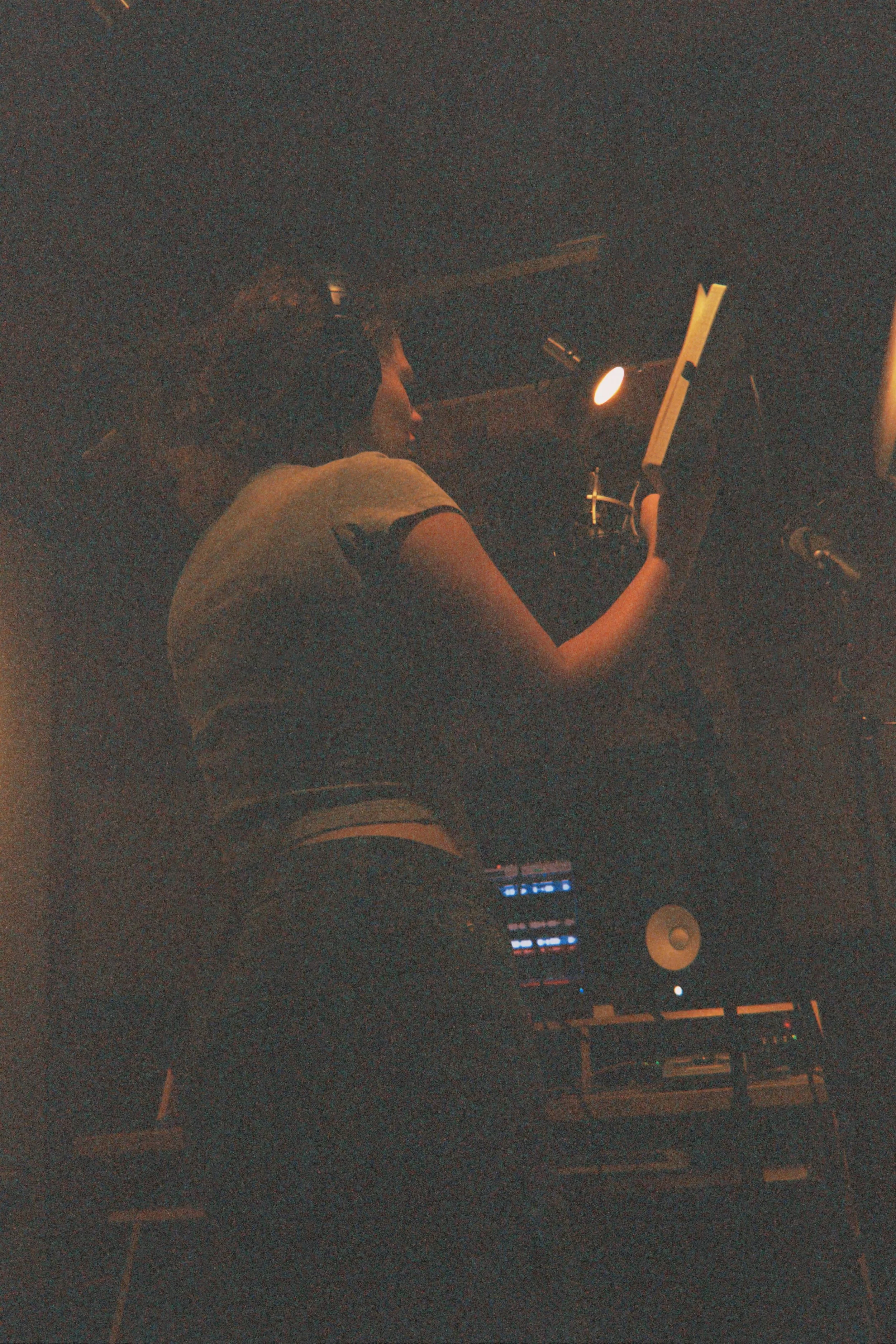 A woman with curly hair is sitting at a music production desk at night, looking at a laptop and audio equipment in a dimly lit room.
