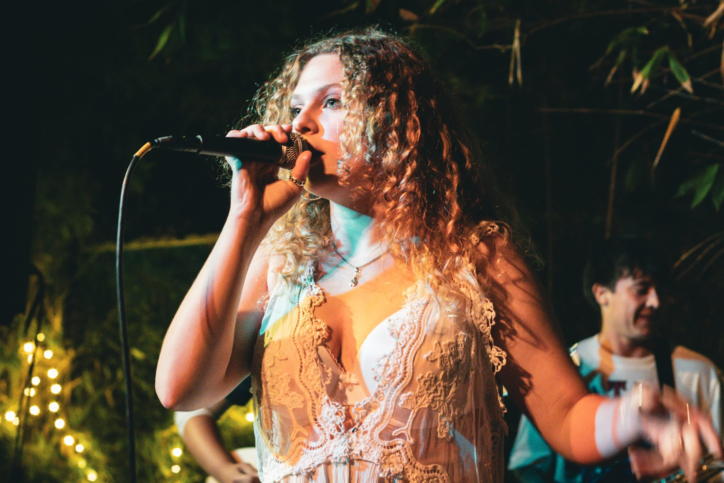 A woman with curly hair holding a microphone, performing at a nighttime outdoor event with string lights in the background.