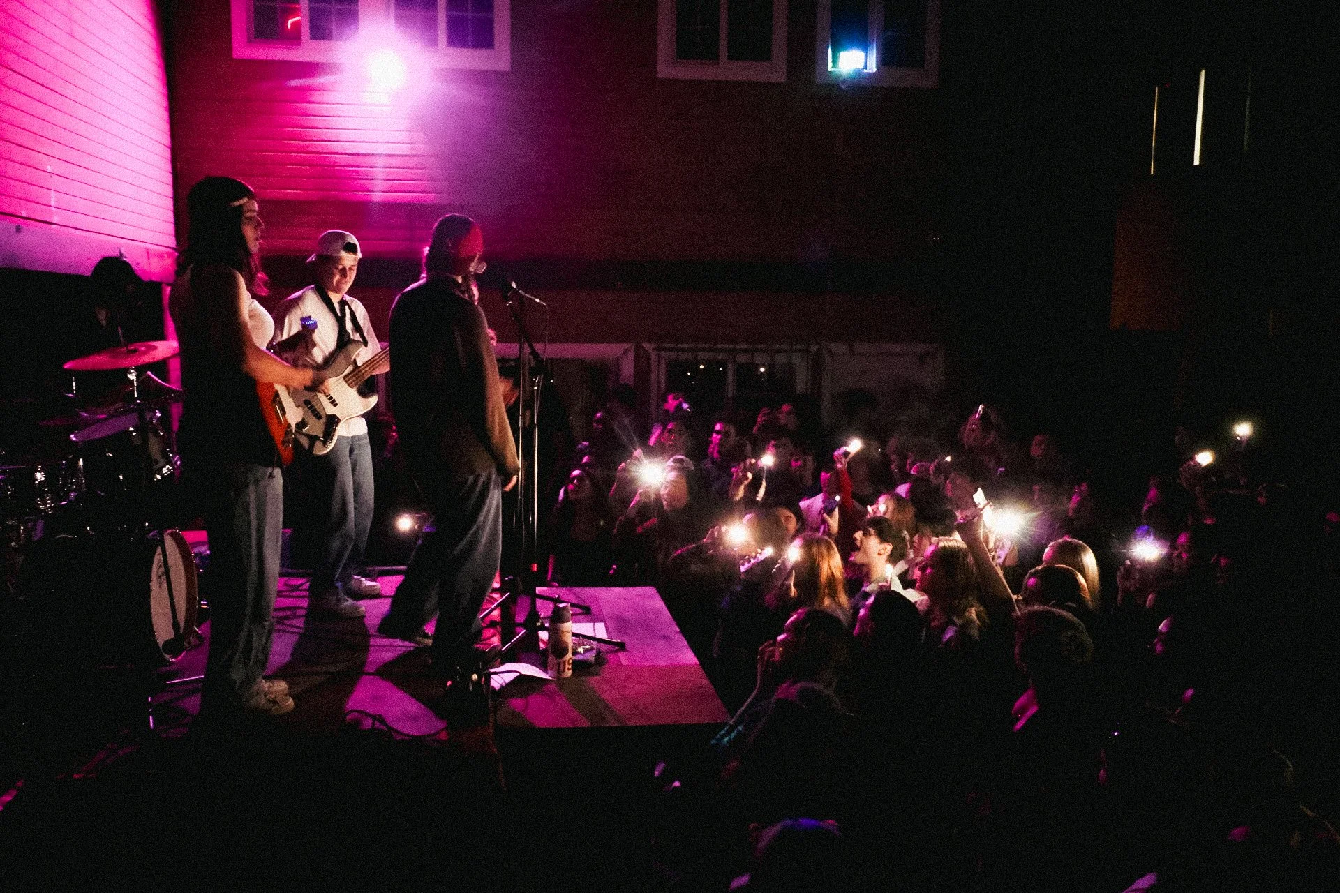 A band performs on stage at a concert or event, with a singer, guitarist, and drummer visible, while the audience holds up phones and lights in the dark.