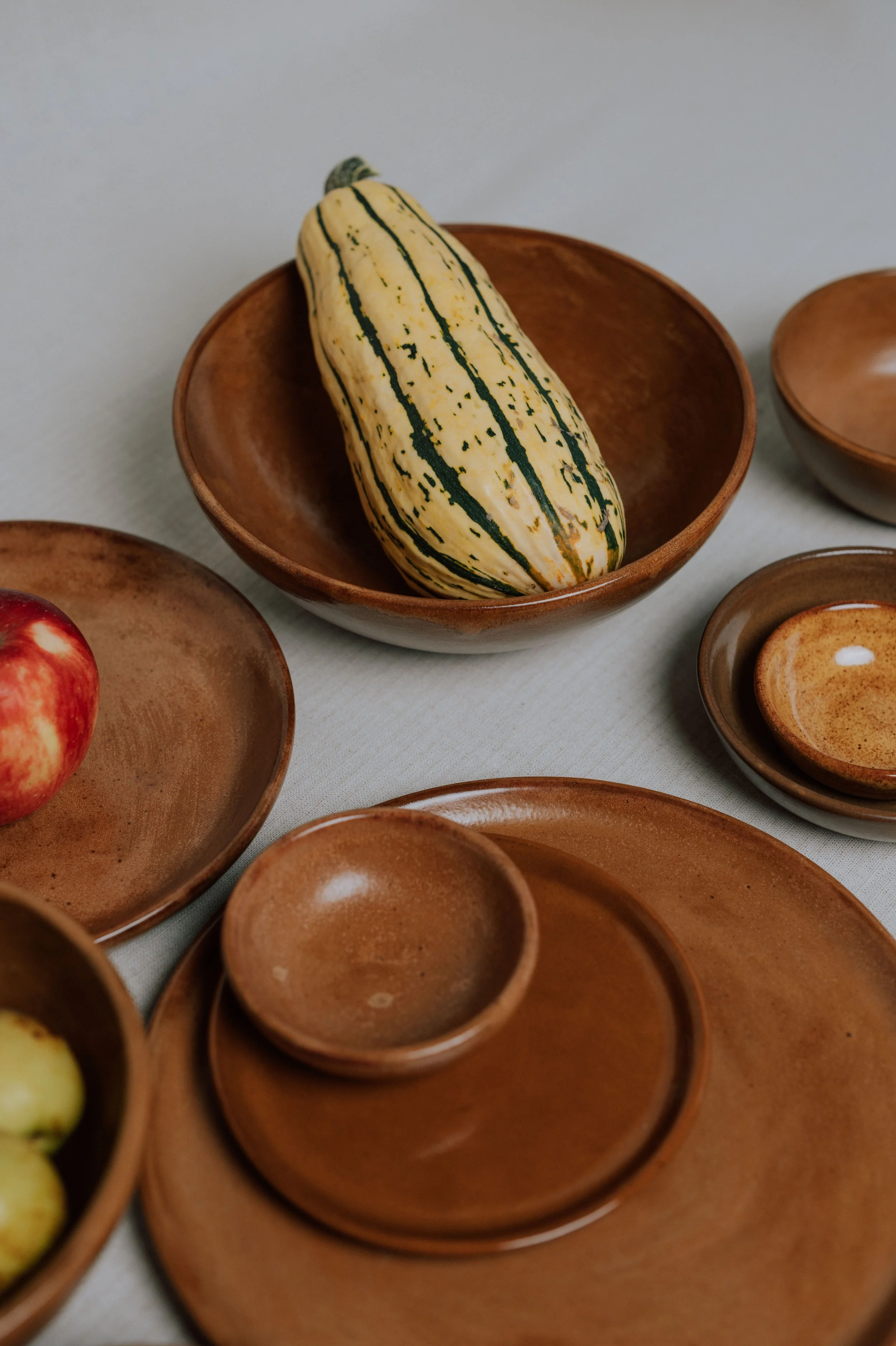 Overhead arrangement of handmade brown ceramic bowls and plates with a striped squash, apples
