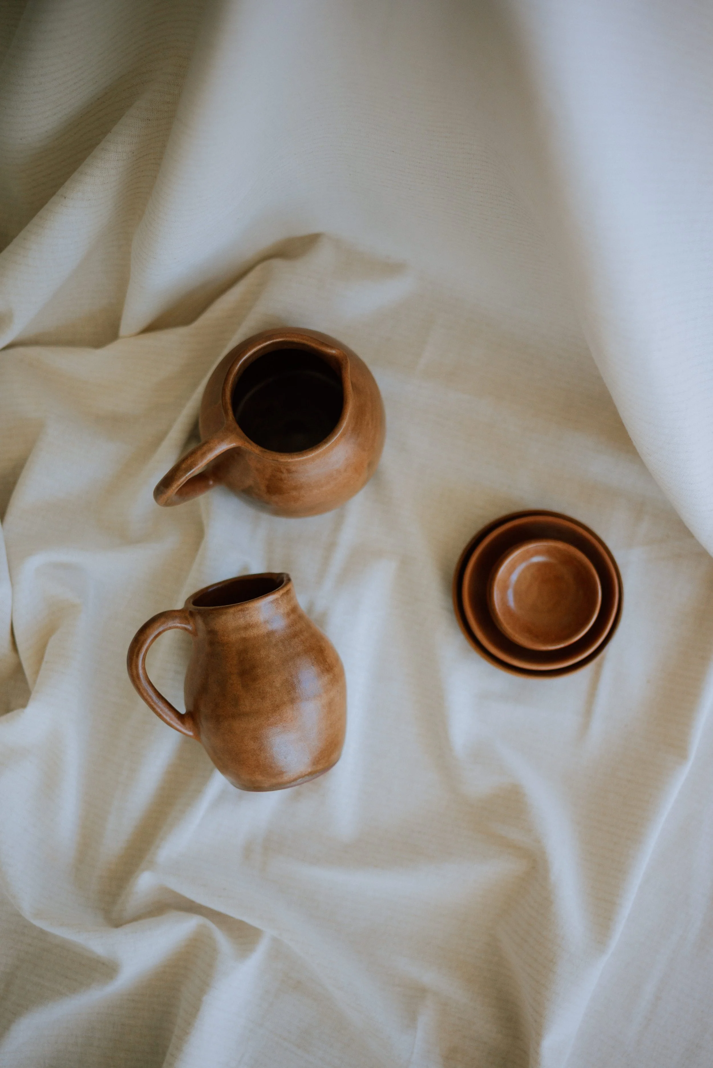 Overhead view of two handmade ceramic pitchers and a stack of small bowls on a soft cream linen backdrop.