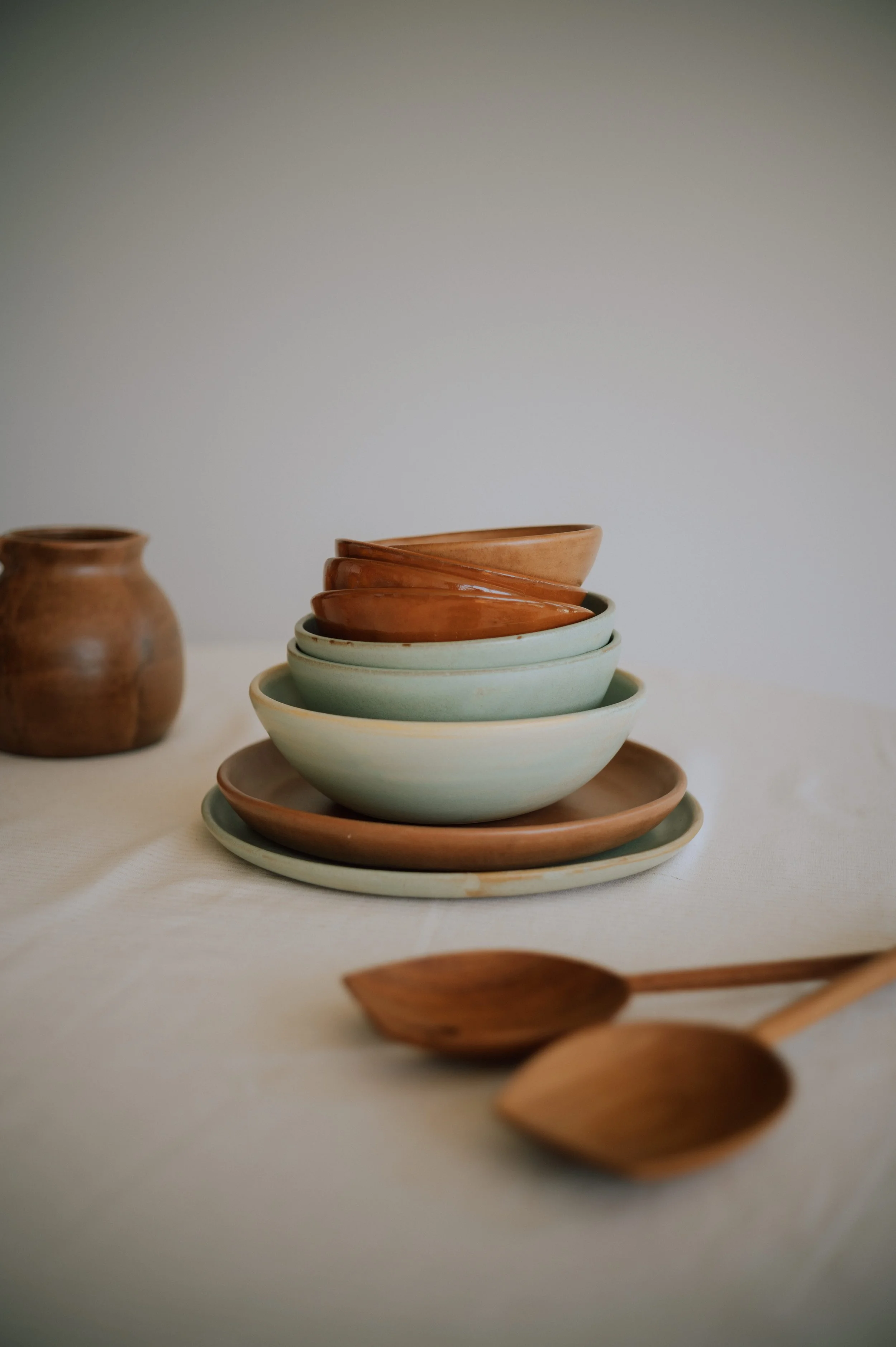 Stack of handmade ceramic bowls and plates in soft sage and terracotta glazes on a linen tablecloth with wooden serving spoons.