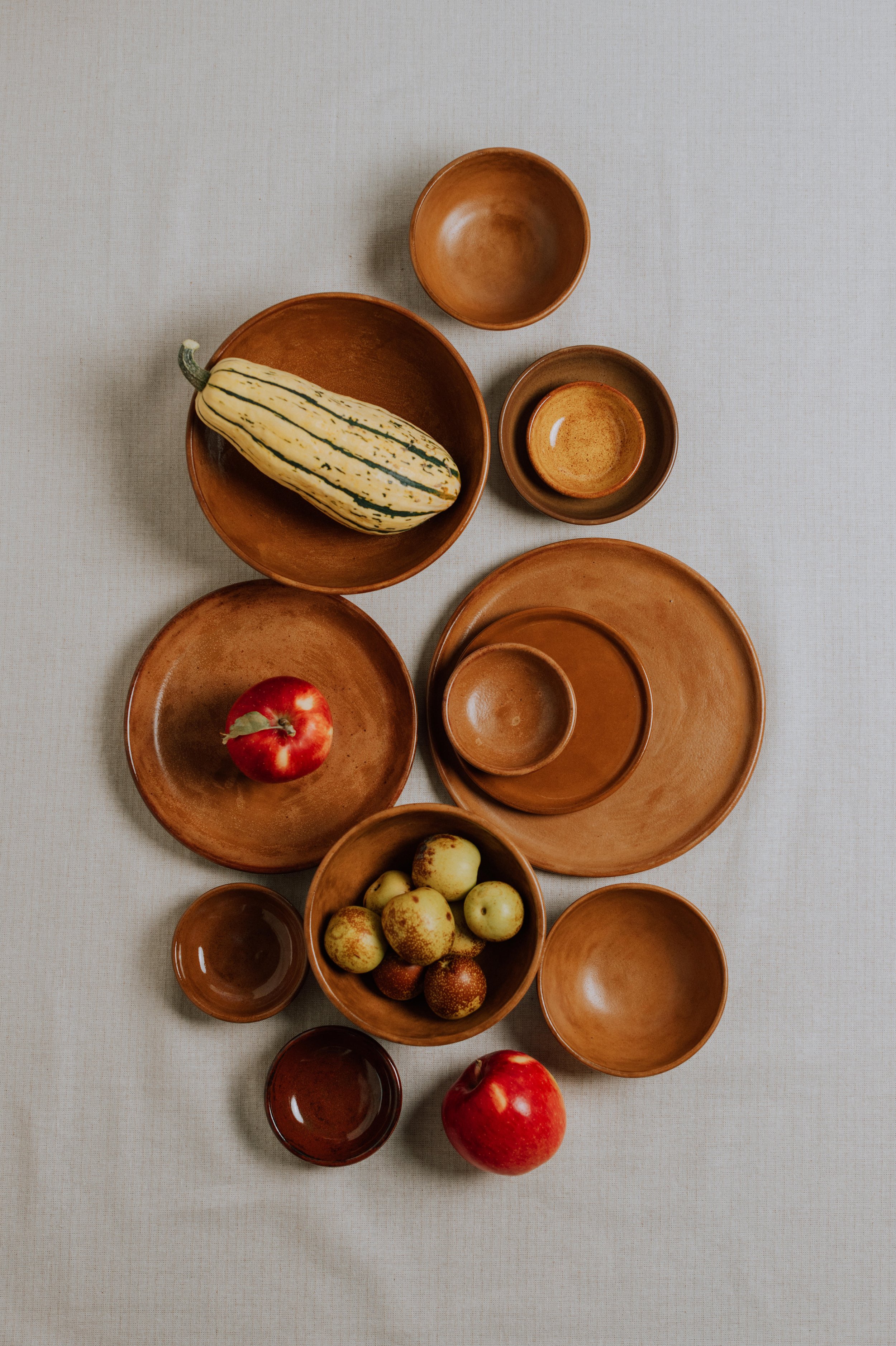 Overhead arrangement of handmade brown ceramic bowls and plates with a striped squash, apples, and small pears