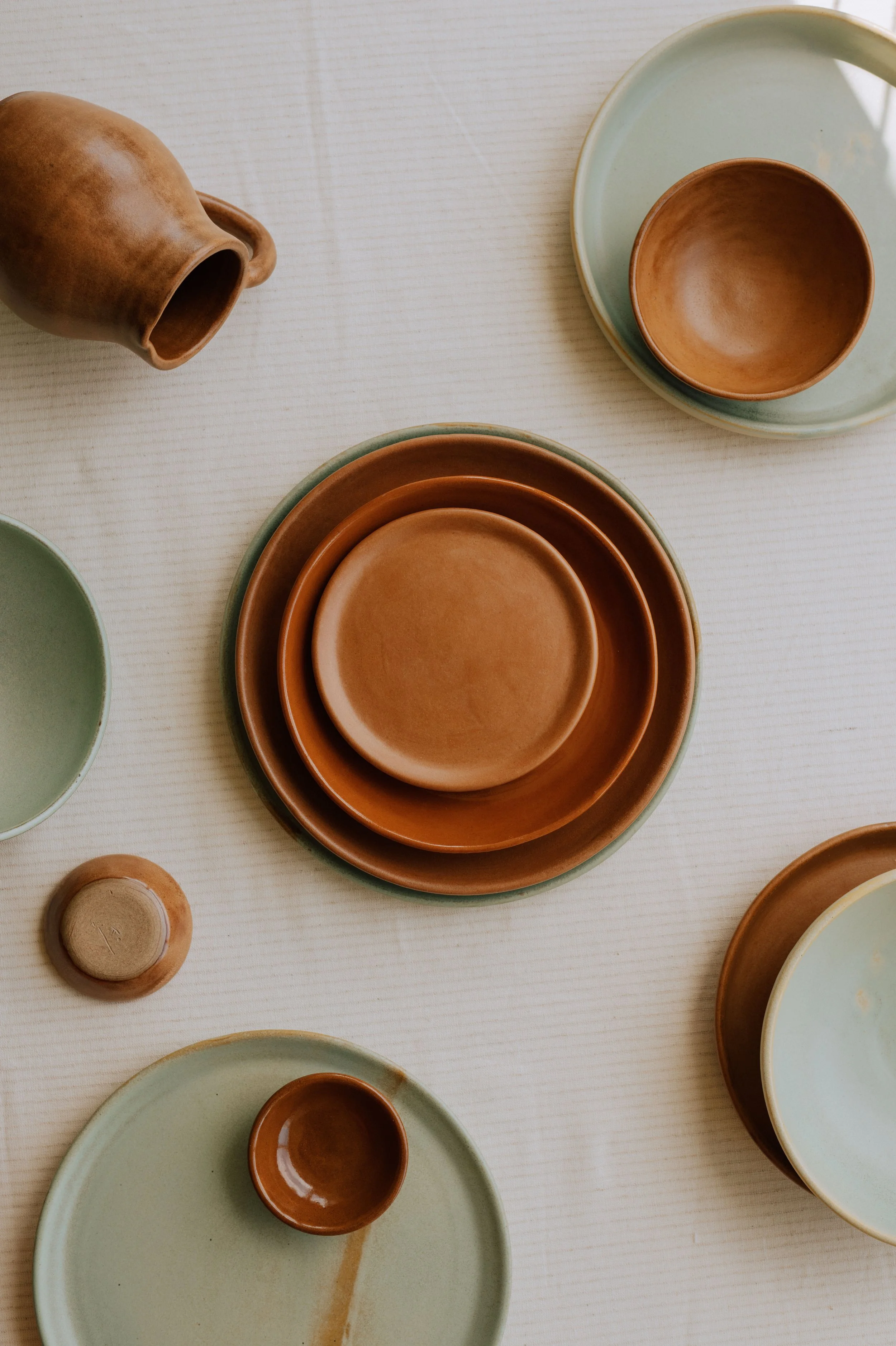 Handmade ceramic dinnerware collection featuring sage green and terracotta plates, bowls, and a pitcher arranged on a natural linen tablecloth.