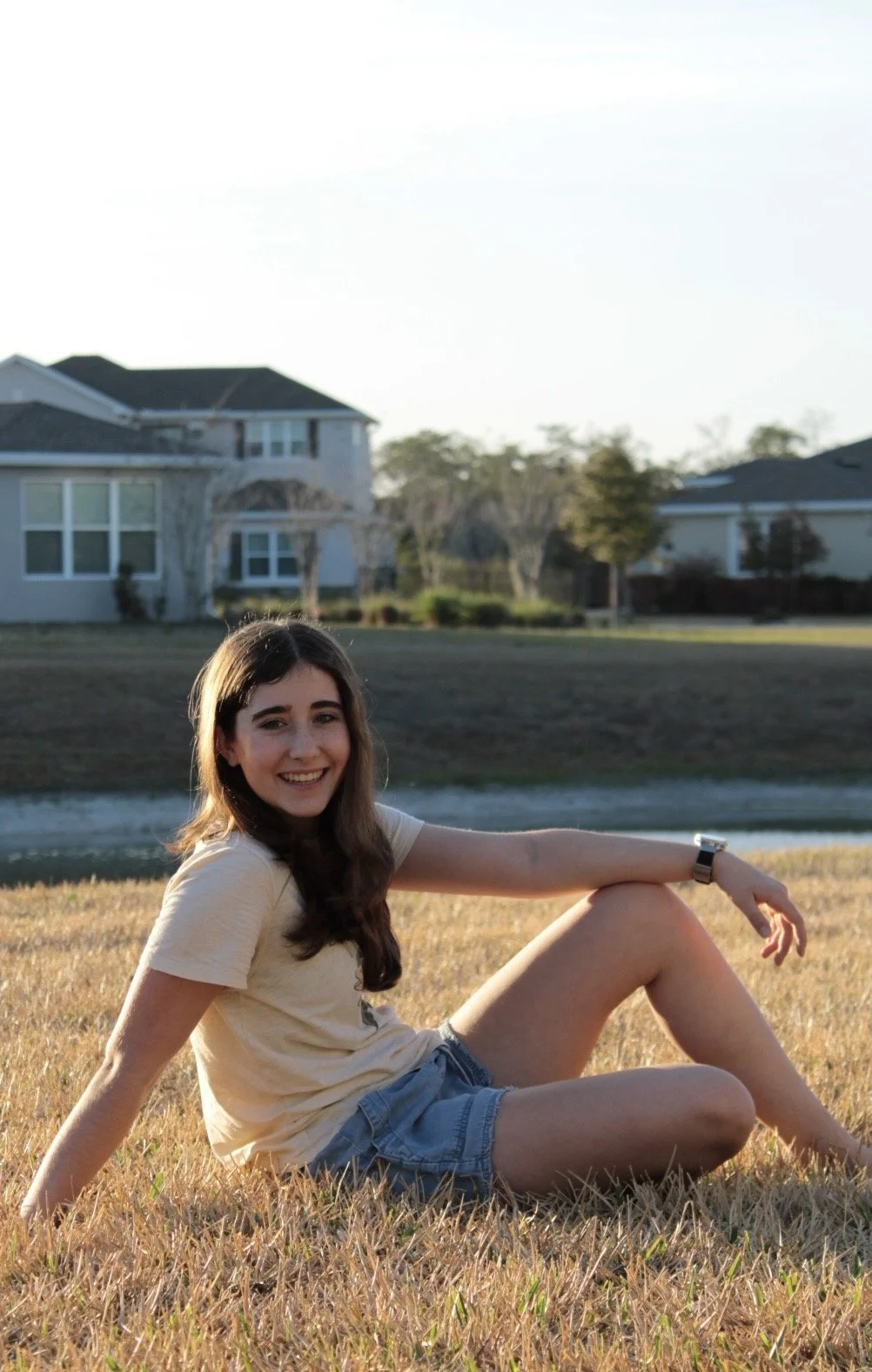 A young woman with brown hair and fair skin sitting on a grassy field near a body of water, smiling at the camera. She is wearing a light-colored T-shirt and denim shorts, with a watch on her left wrist, and there are houses and trees in the background.