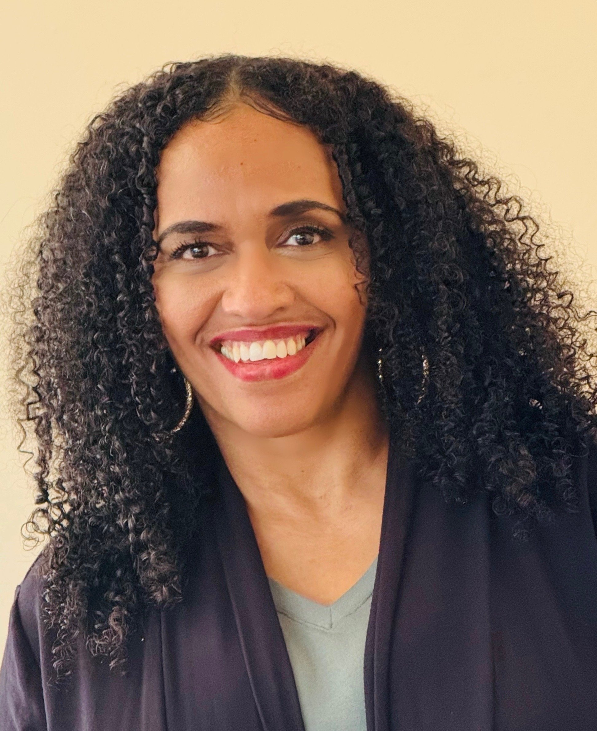 A dark skinned, brown eyed, curly haired woman with hoop earrings smiling