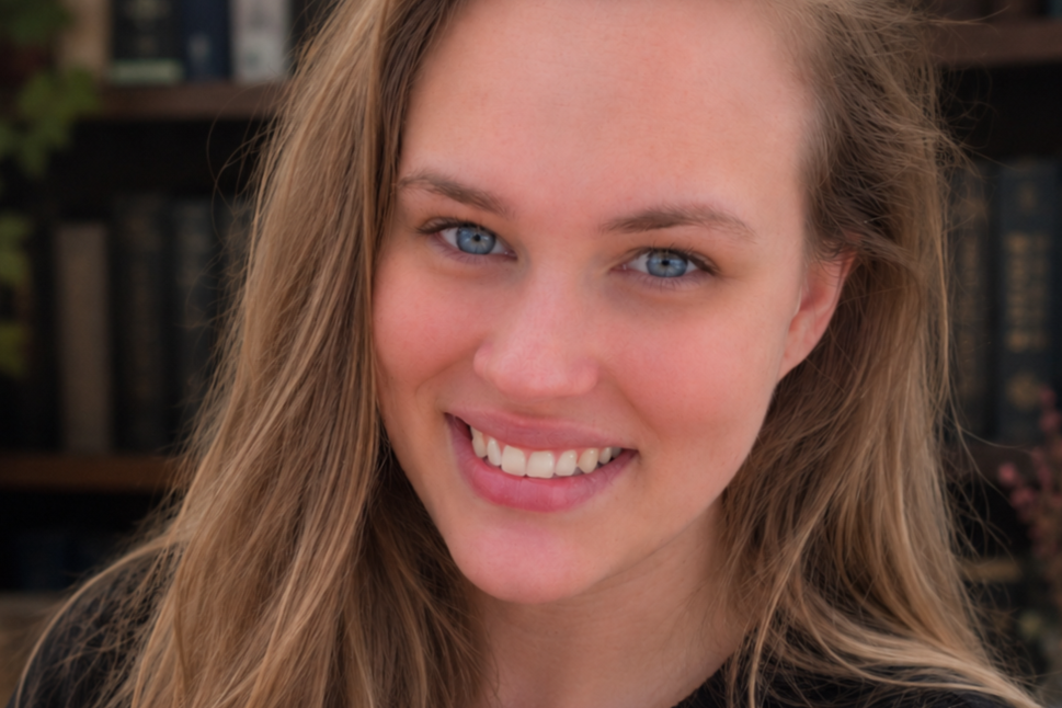 A young woman with long light brown hair, blue eyes, and a bright smile, in front of a blurred bookshelf background.