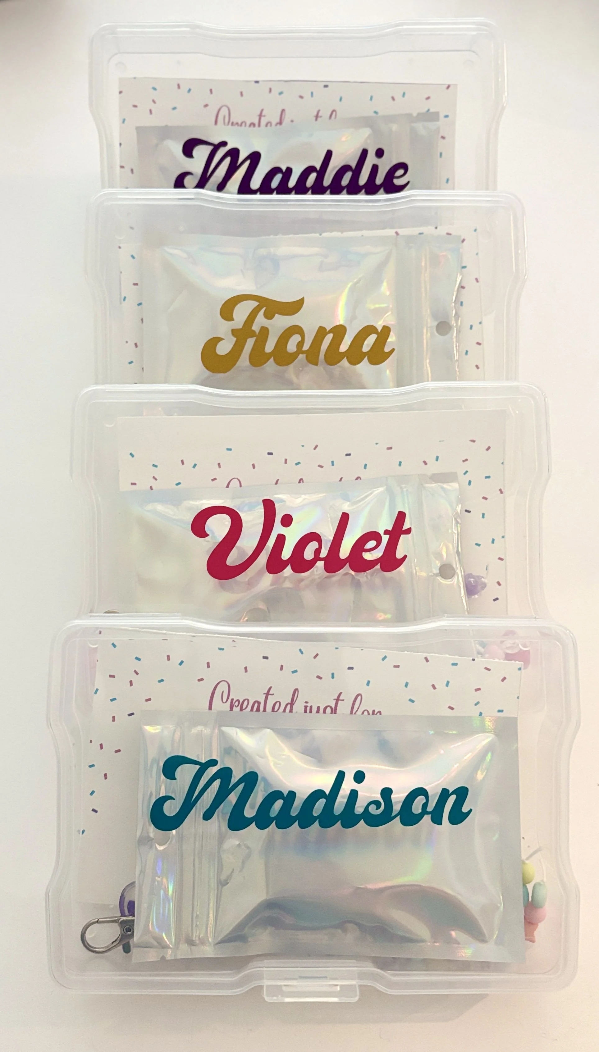 Clear plastic organizer with four compartments labeled in colorful text: Maddie, Fiona, Violet, Madison, containing various items.