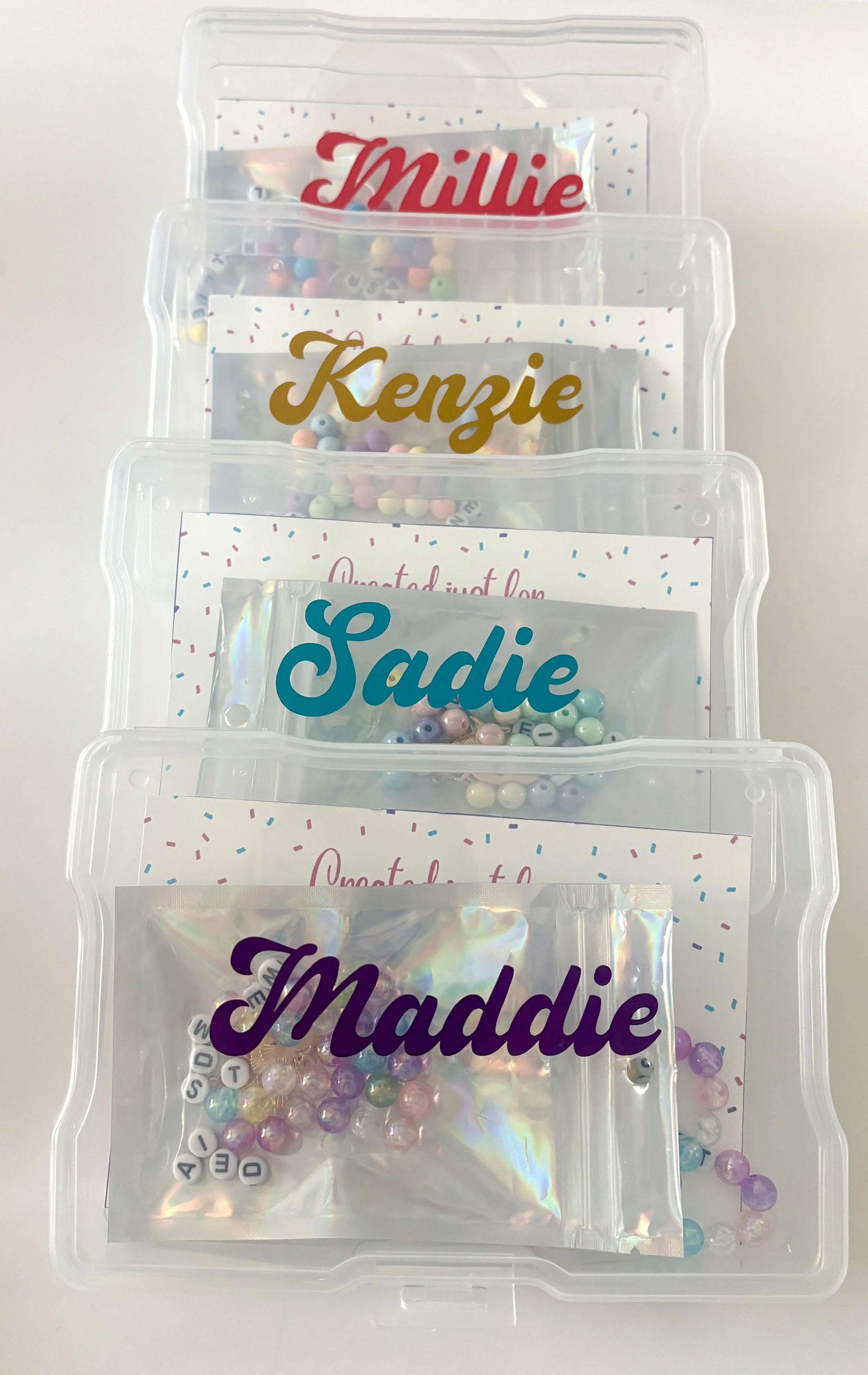 Clear plastic storage container with four smaller sealed compartments, each labeled with a different name in colorful cursive font, containing pastel-colored beads and lettered alphabet beads for jewelry making.