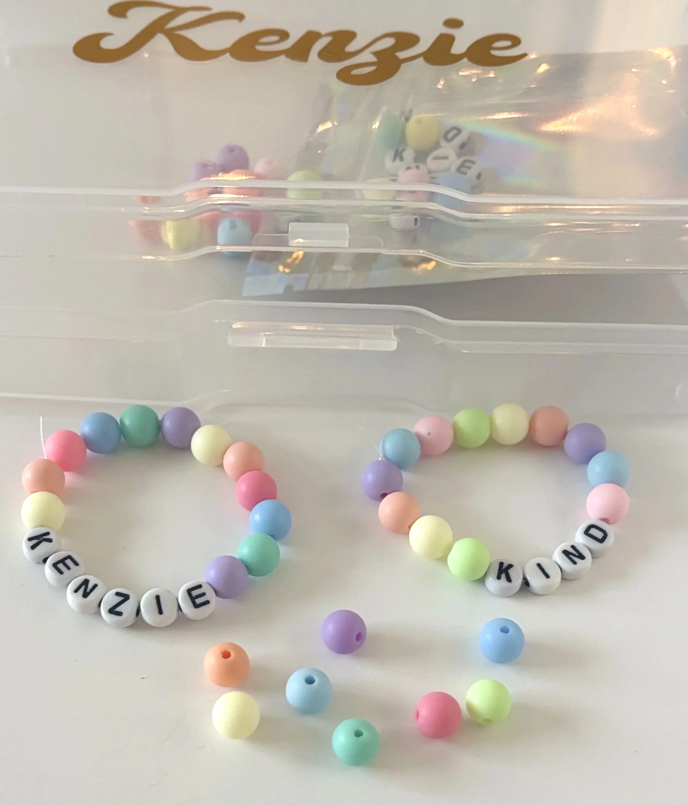 Colorful beaded necklace spelling out 'KINDNESS' with pastel-colored beads, placed on a white surface with a clear plastic box labeled 'Kensie' in the background.