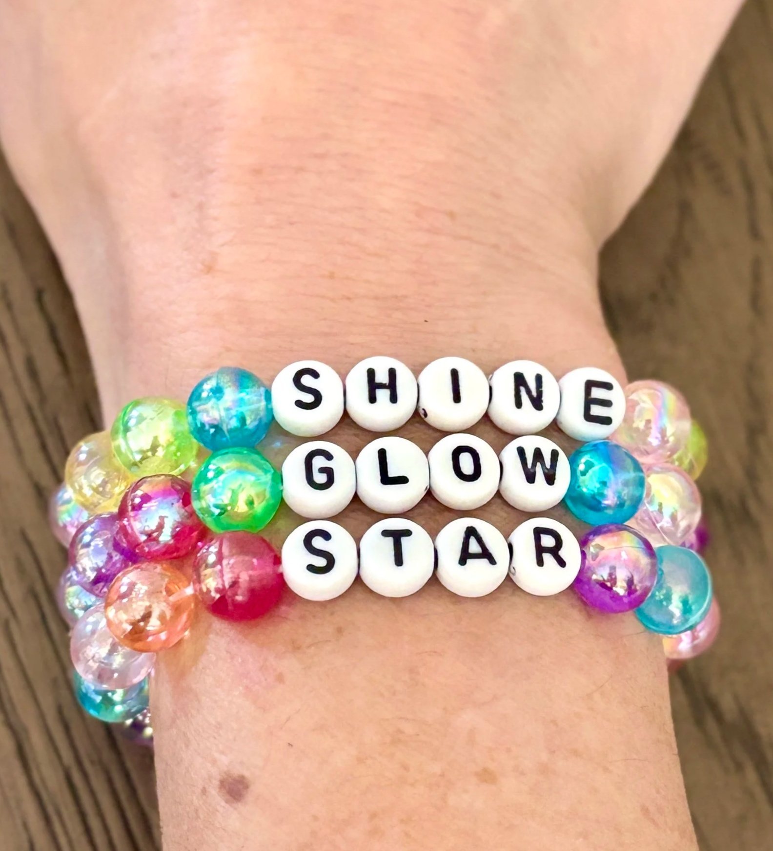 Close-up of a person's arm adorned with colorful beaded bracelets and letter beads spelling out words like 'SHINE,' 'GLOW,' and 'STARS.'