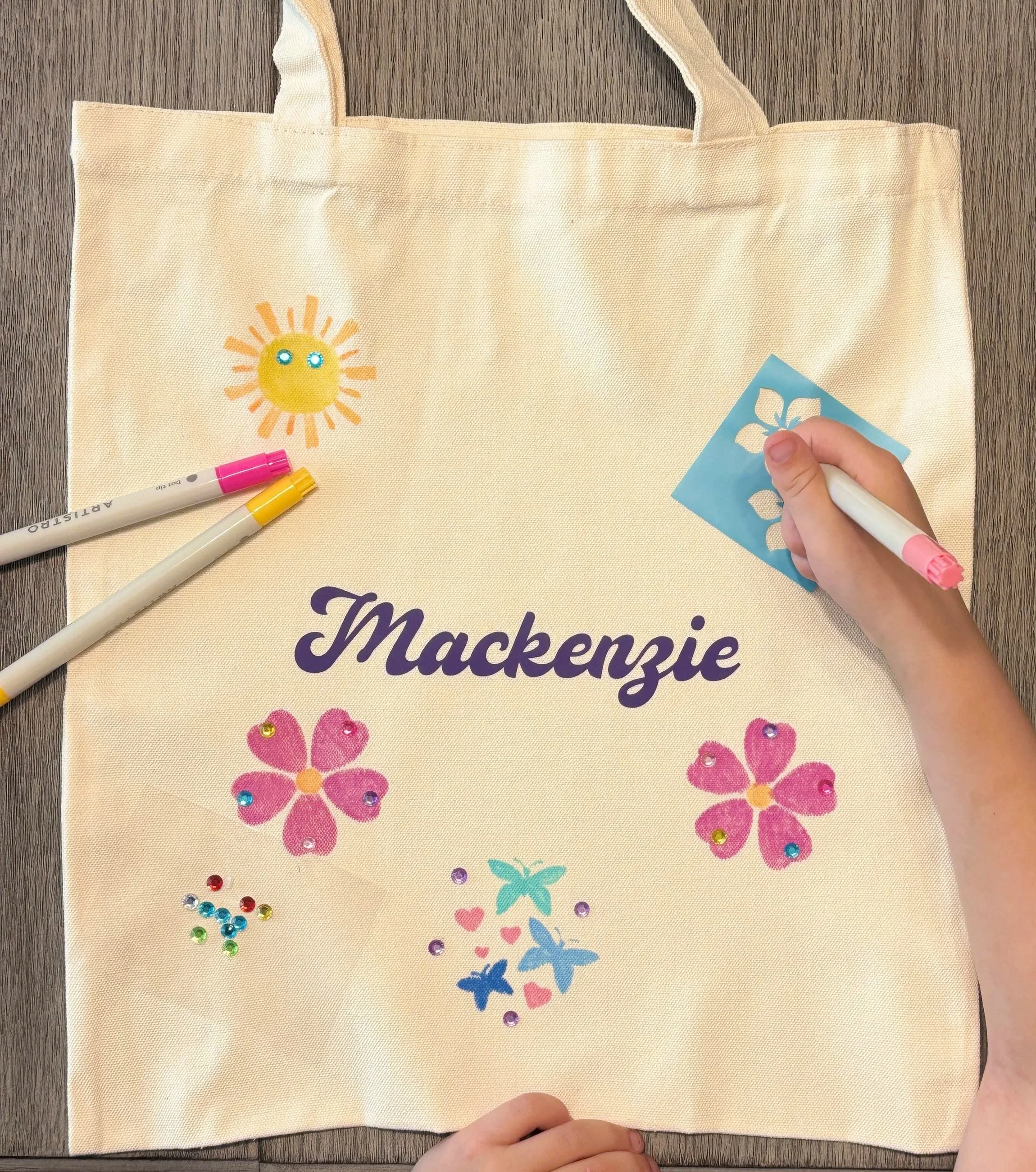 A beige tote bag on a wooden surface decorated with colorful stickers of butterflies, flowers, and a sun.