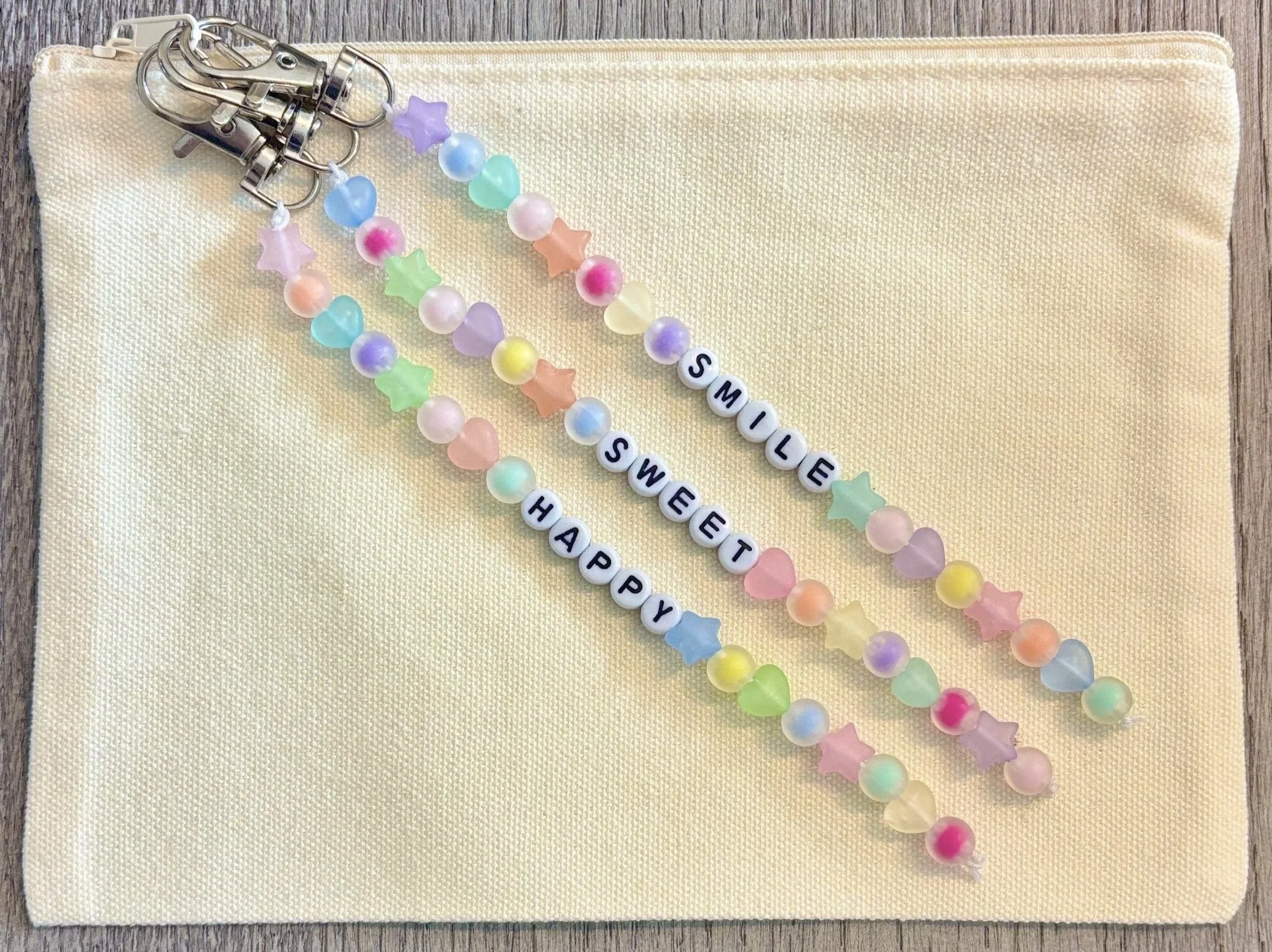 Colorful beaded bracelets with star and heart shapes, spell out "SMILE," and decorative beads including pearls. They are placed on a cream fabric pouch on a wooden surface.