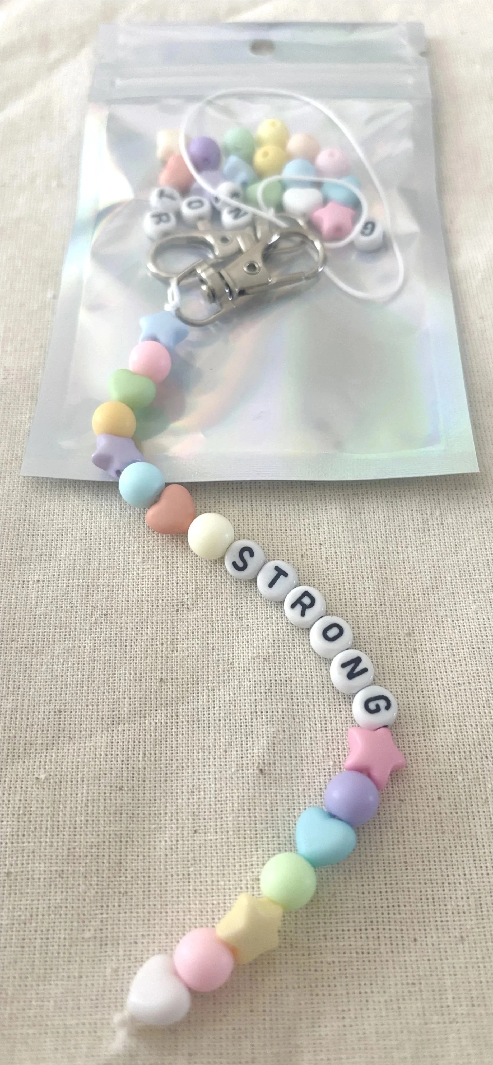 A pastel-colored beaded necklace spelling out the word 'STRONG' with alphabet beads, along with a bag of assorted pastel beads and a metal clasp.