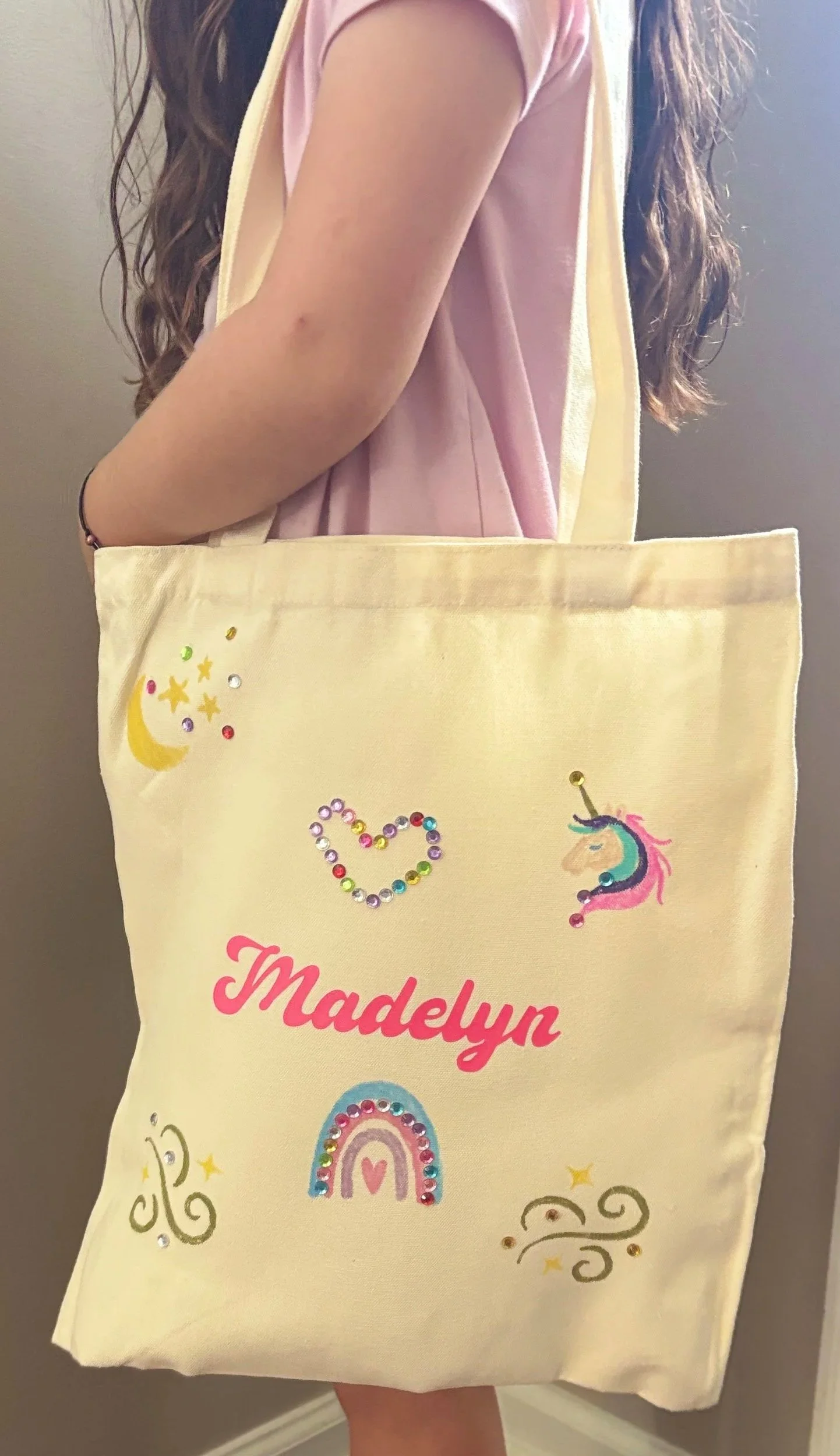 A person holding a yellow tote bag with colorful unicorn, rainbow, moon, and star decorations. The person is wearing a pink short-sleeved shirt and has long wavy brown hair.