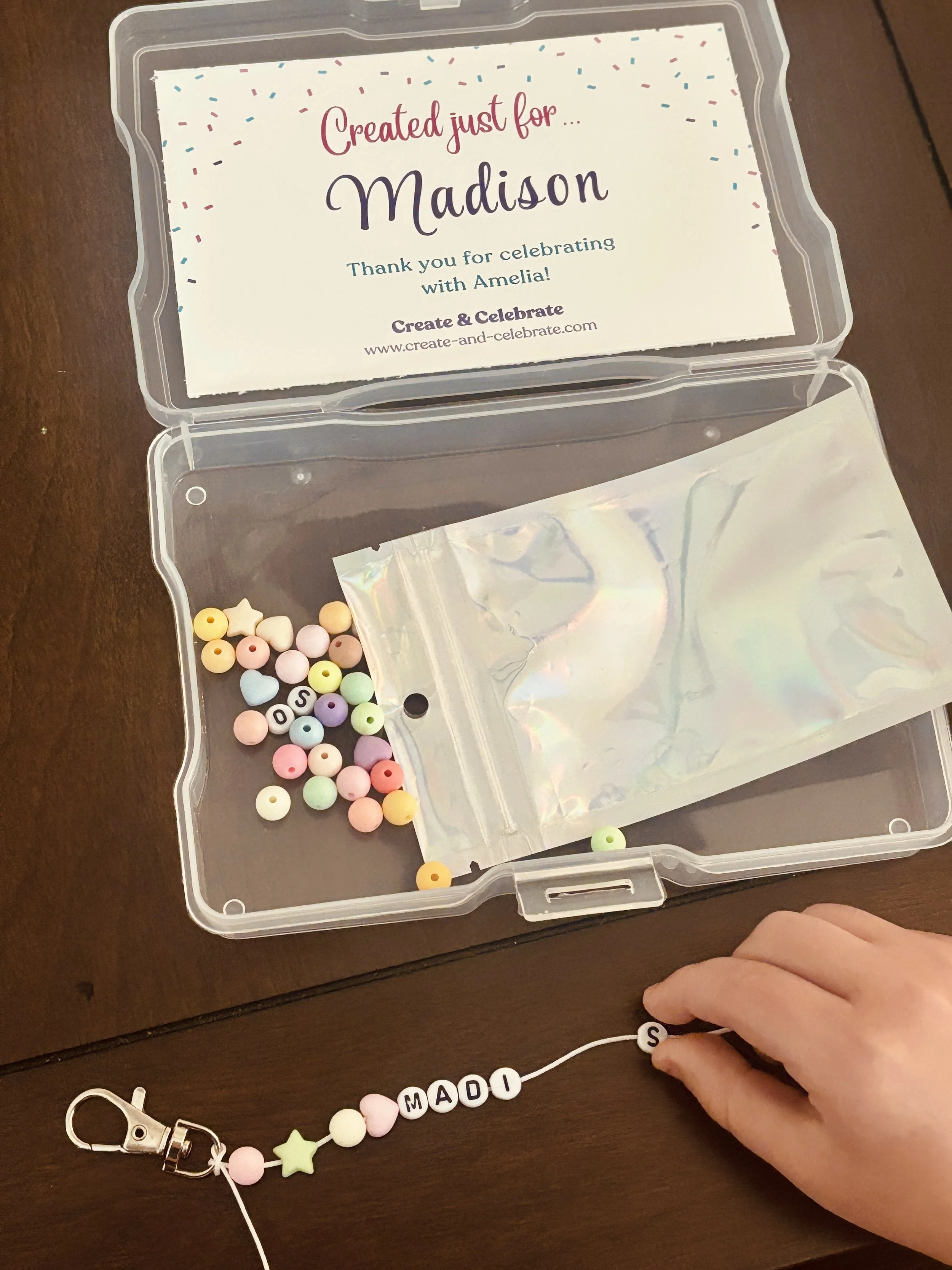 A plastic container with a thank you note for a person named Madison, decorated with colorful confetti. A hand is holding a beaded name tag spelling 'MADI' with a clip attached, and there are pastel-colored beads and a star-shaped bead inside the con