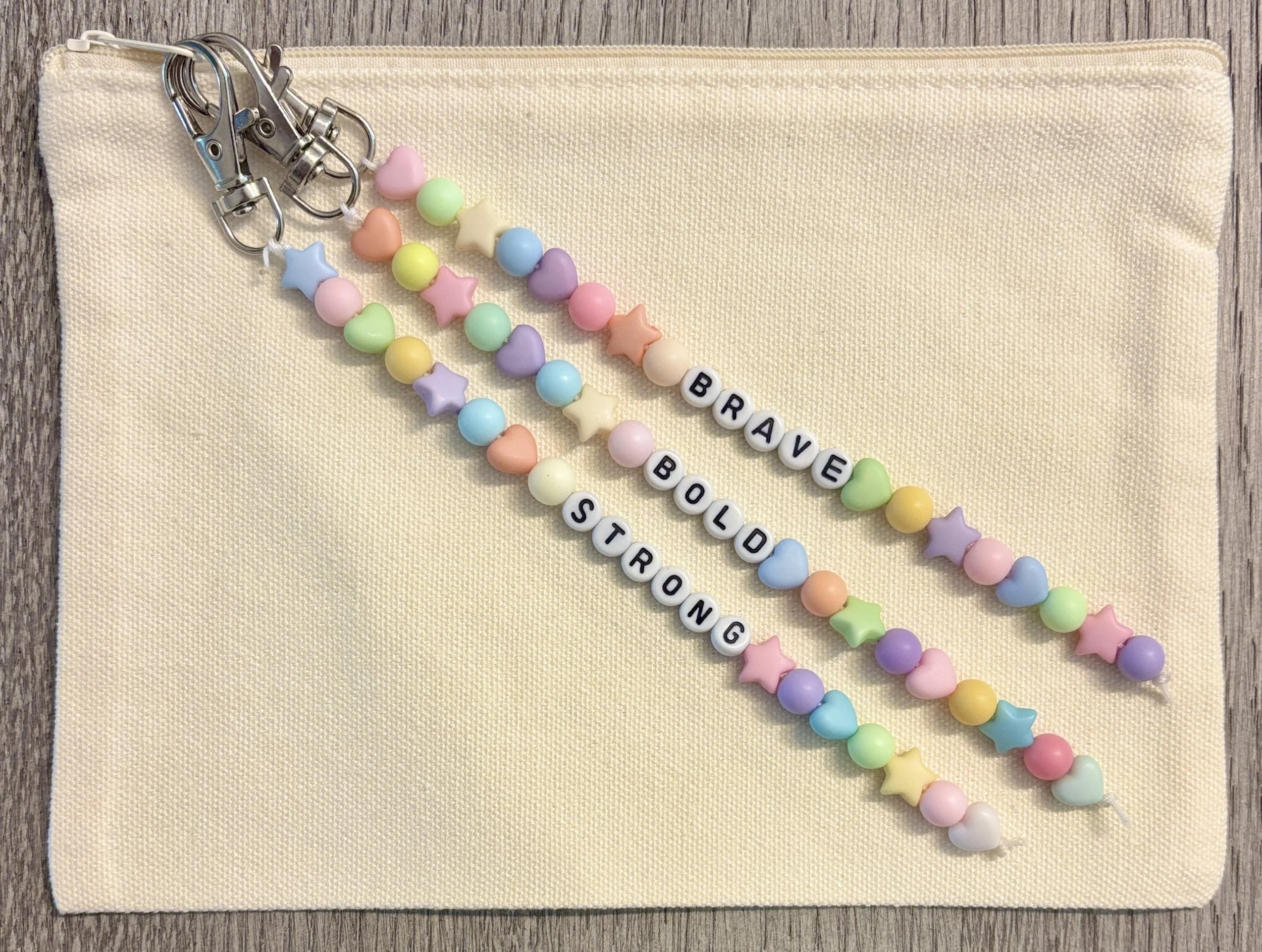 Colorful beaded keychain with pastel hearts, stars, and round beads that spell out "BRAVE BLOSSOM" on a beige fabric pouch, on a wooden surface.