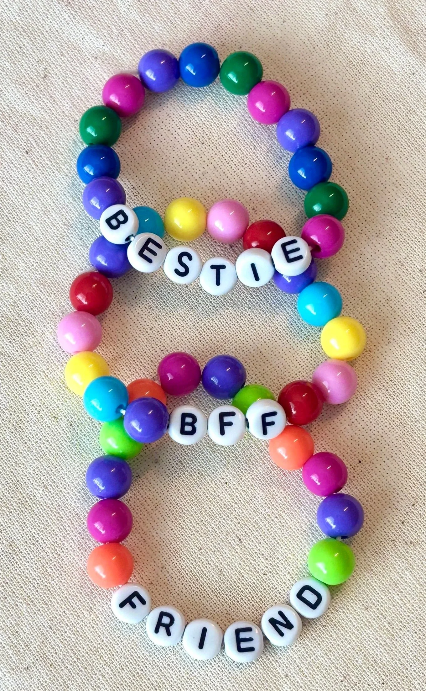 Colorful beaded bracelets with letter beads spelling out "BESTIE," "BFF," and "FRIEND" on a light fabric background.