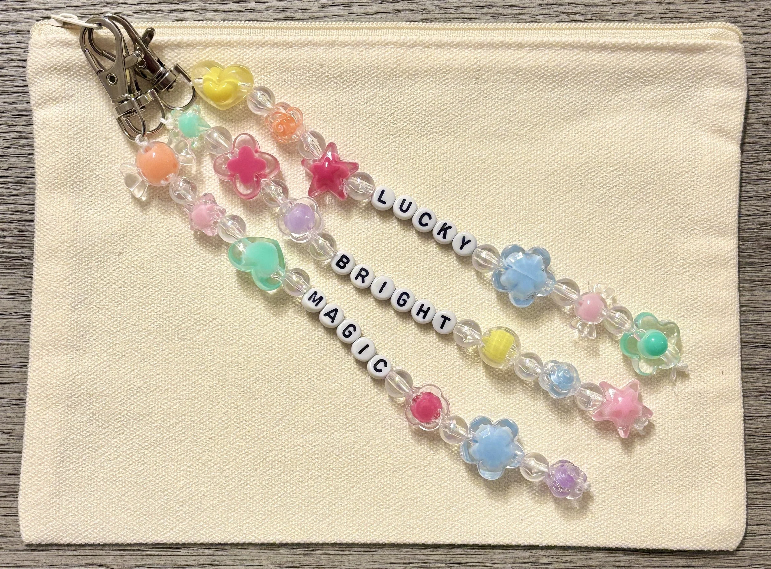 Colorful beaded bracelets with pastel hearts, stars, and letter beads spelling out 'LUCKY BRIGHT MAGICAL' on a cream fabric background.