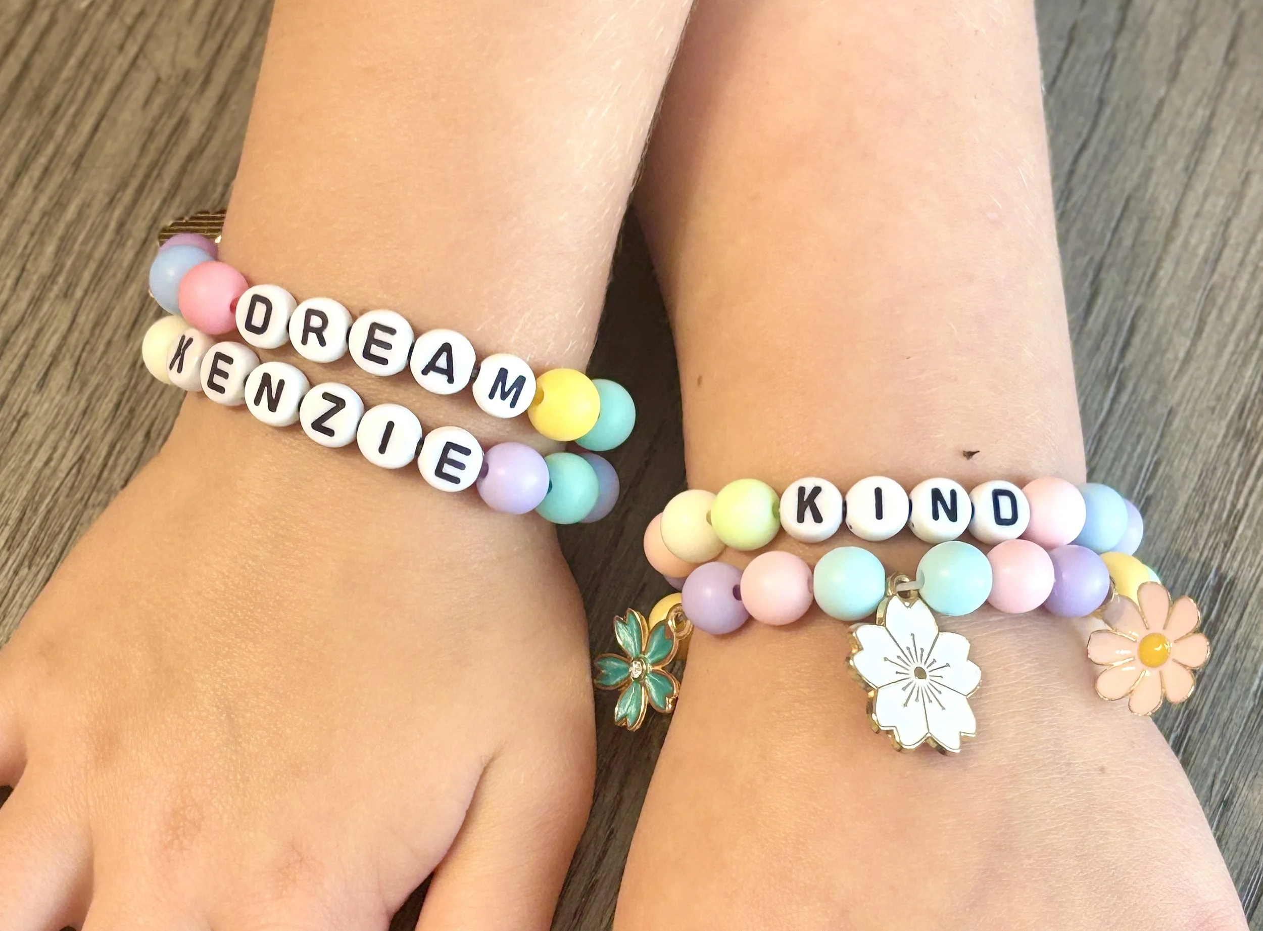 Close-up of a person's wrist wearing multiple colorful beaded bracelets with letter beads spelling 'DREAM' and 'KIND', and floral charms, on a wooden surface.