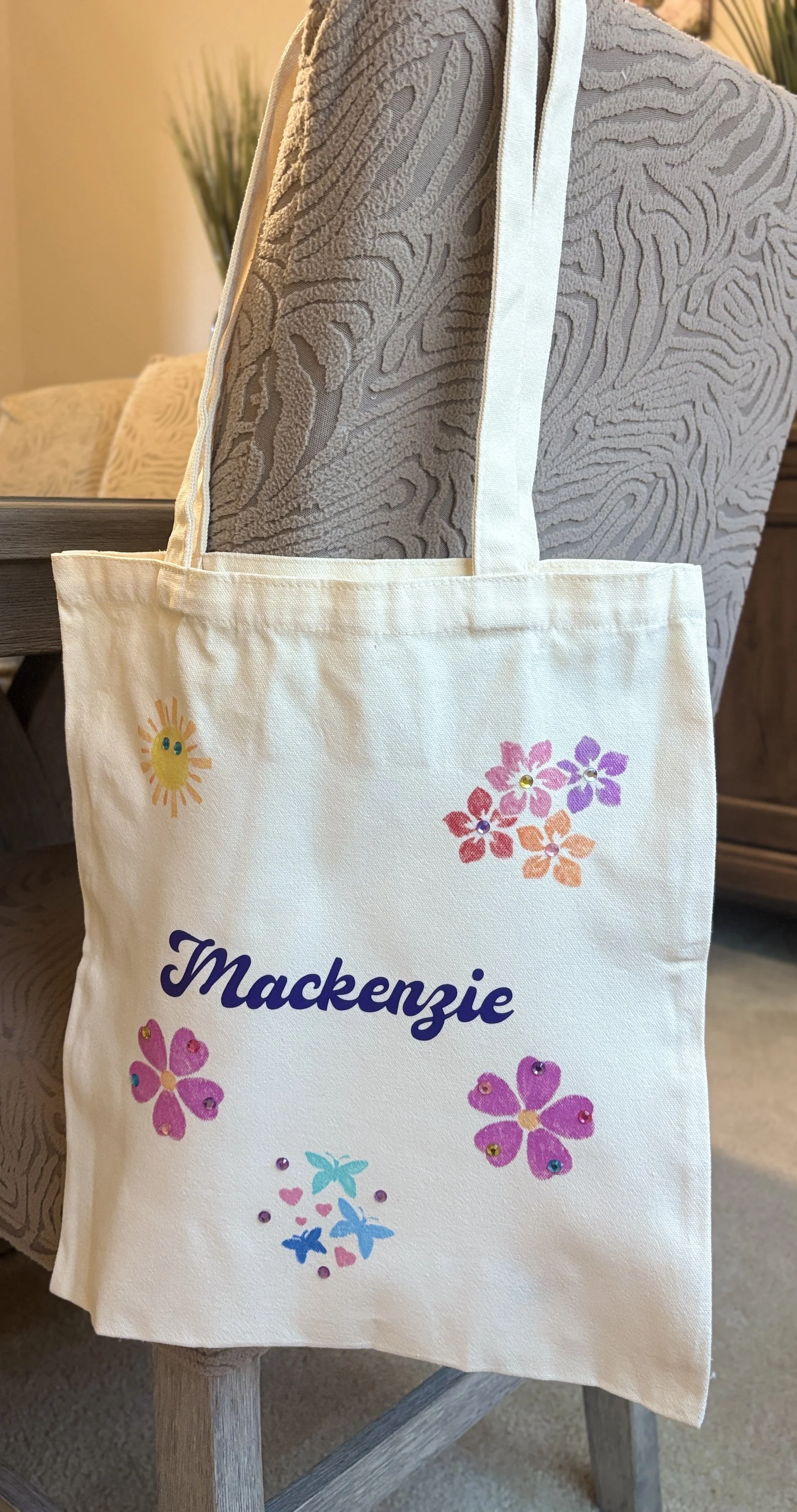 A white tote bag hanging on the back of a chair. The bag has colorful flower and butterfly decorations and the name "Mackenzie" written on it.