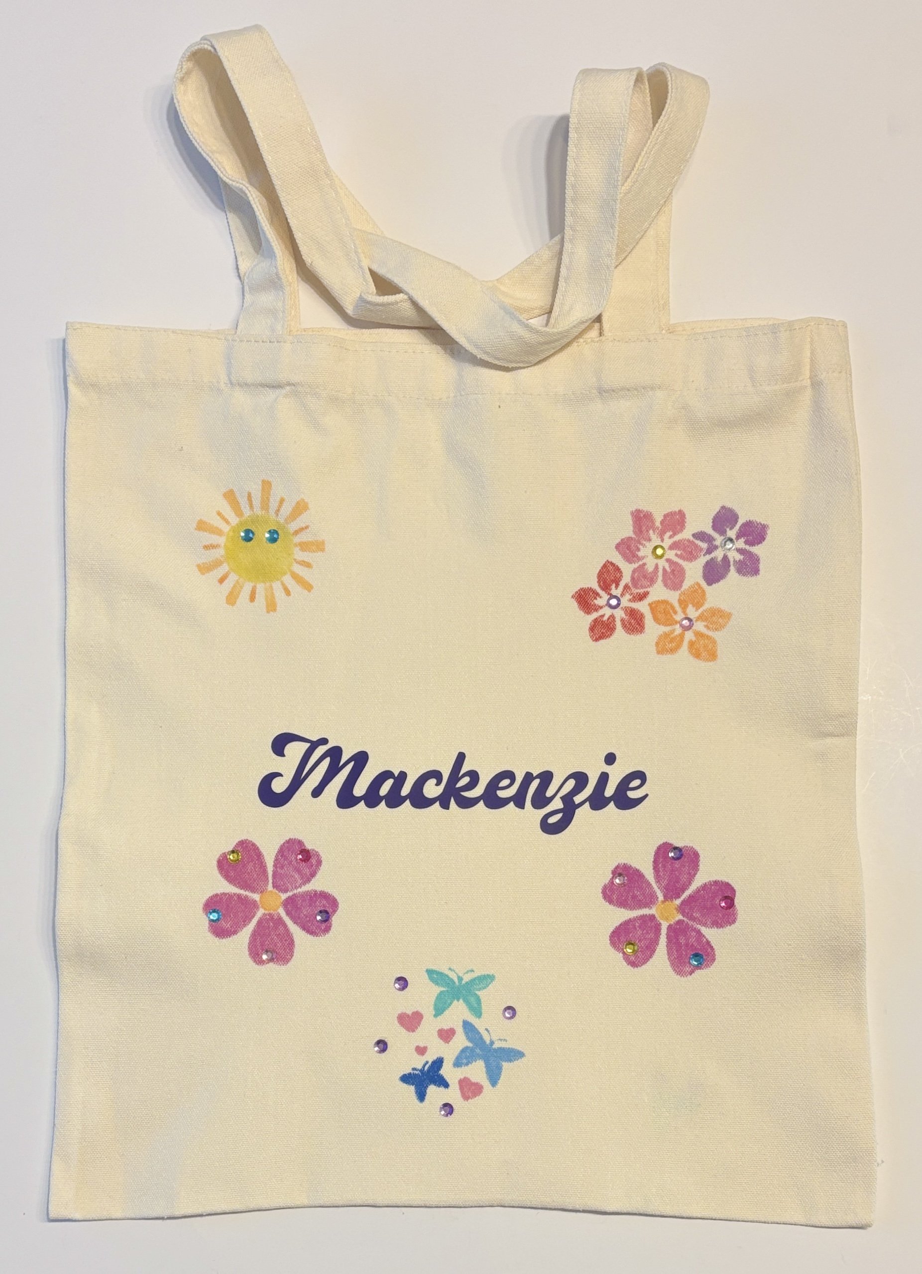 A beige tote bag with colorful flower, butterfly, sun, and heart designs and the name 'Mackenzie' printed in purple.