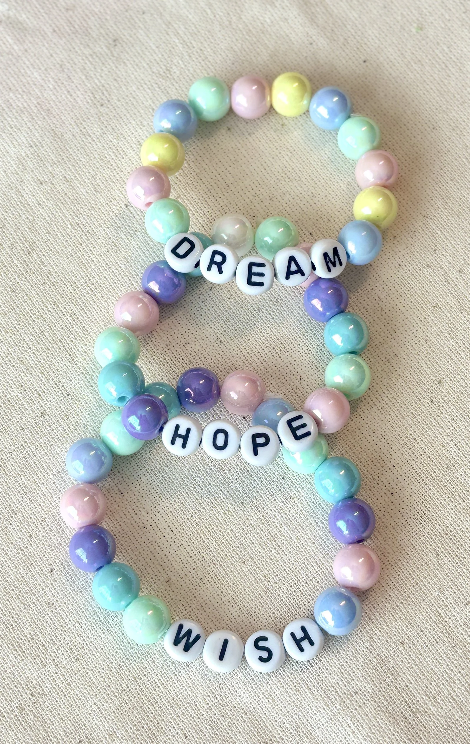 Colorful beaded bracelets with word beads spelling out "DREAM," "HOPE," and "WISH" on a beige fabric background.