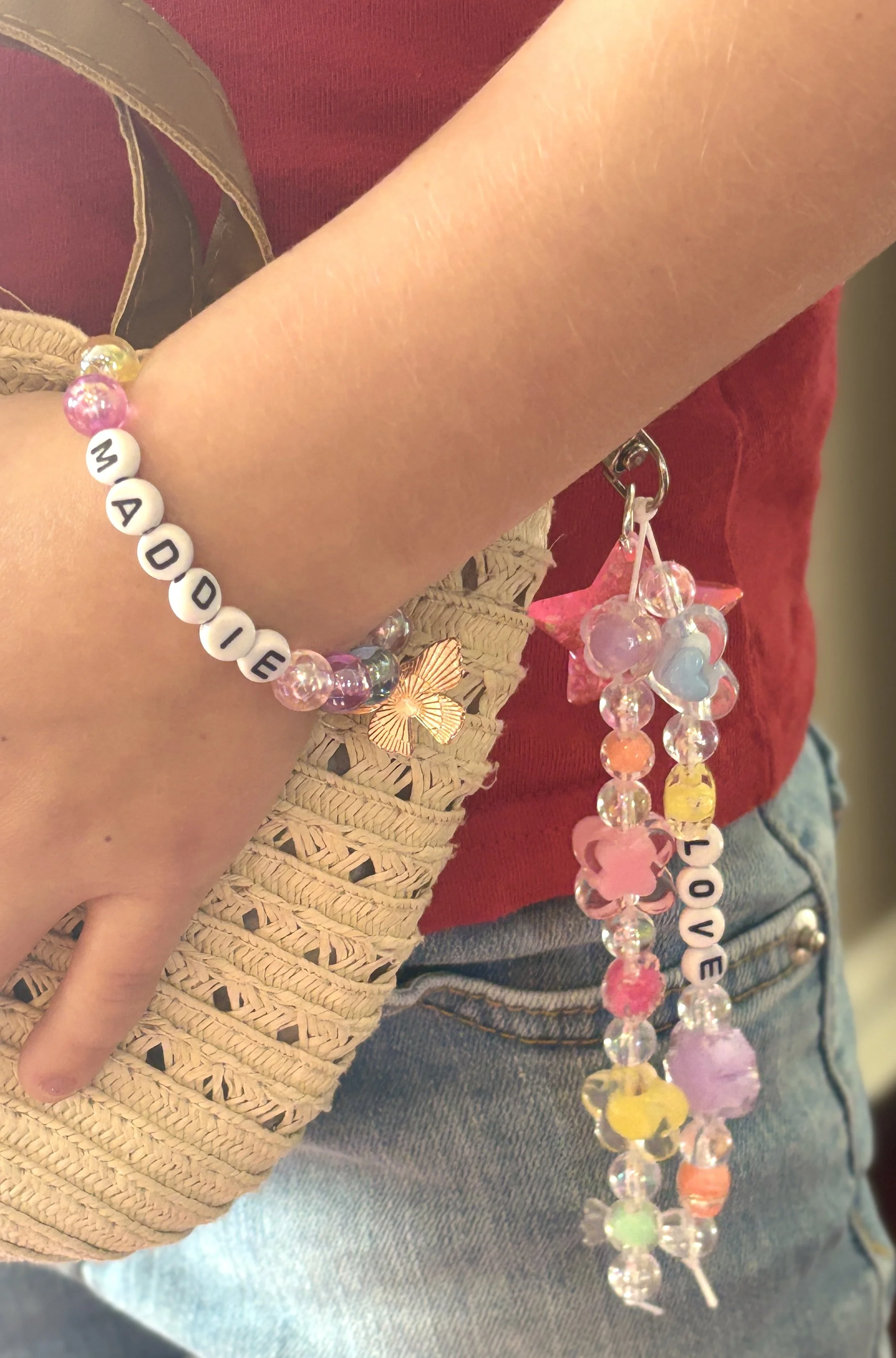 A person's arm and hand are shown, wearing a bracelet with multicolored beads spelling out 'MADDIE' and a star-shaped pink bead. The person has some light scratches on their skin. They are also holding a beaded necklace with multicolored beads, some 
