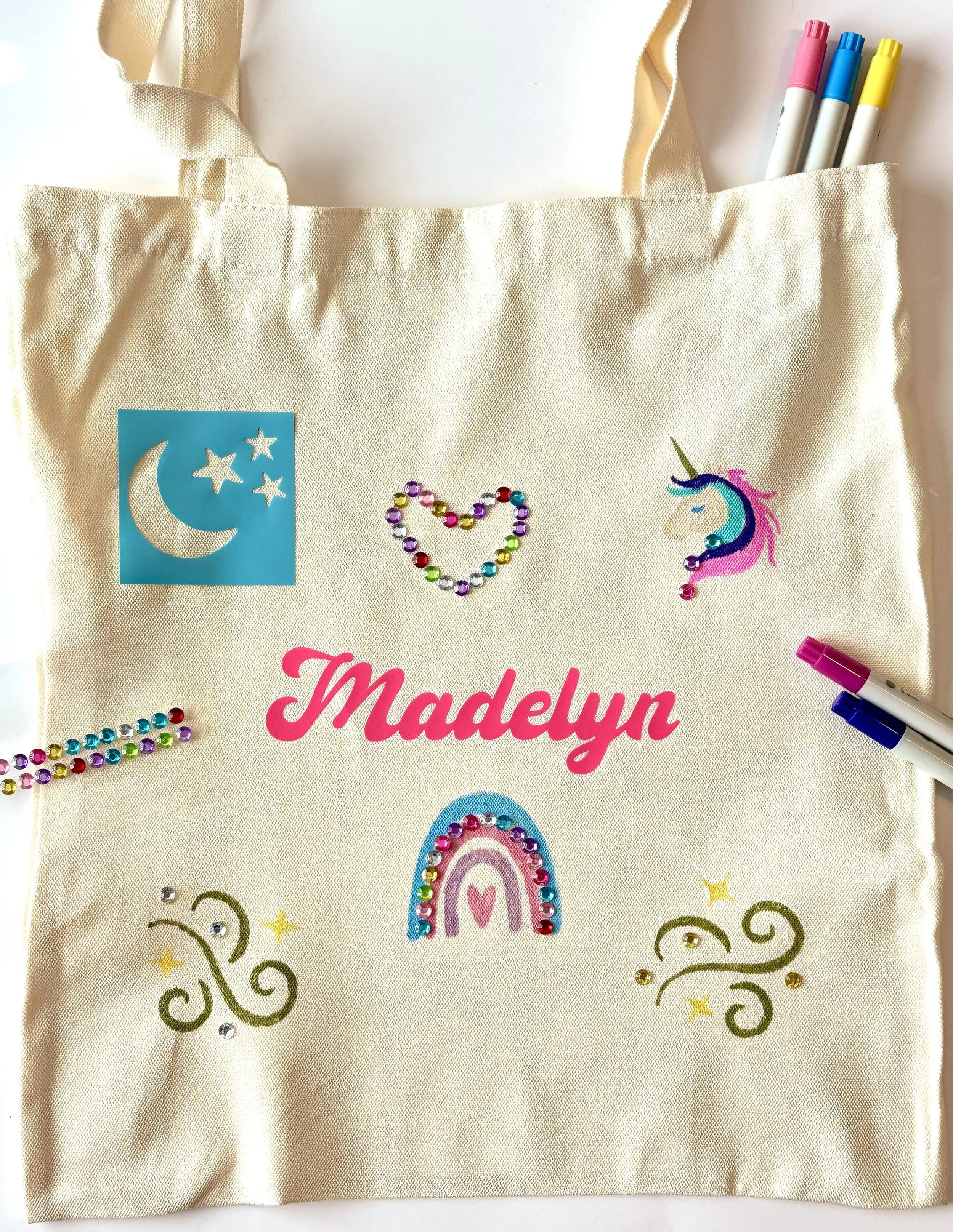 A beige canvas tote bag with colorful decorations and the name "Madelyn" in pink text. The bag has a rainbow, a unicorn, a moon and star sticker, gold swirls, and rhinestones. There are four markers inside the bag.