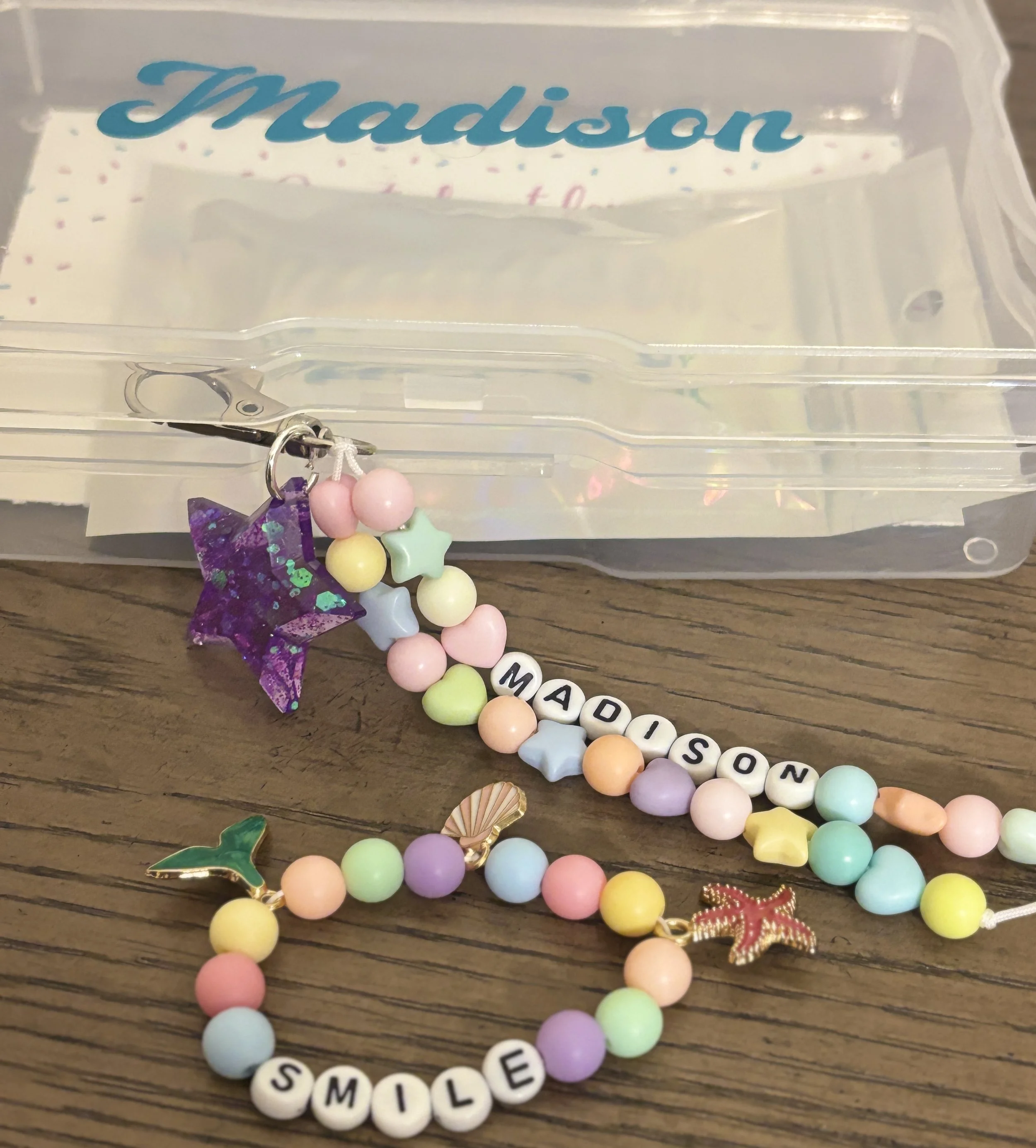Clear plastic box with the name "Madison" on the inside lid. Several colorful bead bracelets, rhinestone star and butterfly charms, and letter beads spelling "SMILE" are arranged in front of the box on a wooden surface.