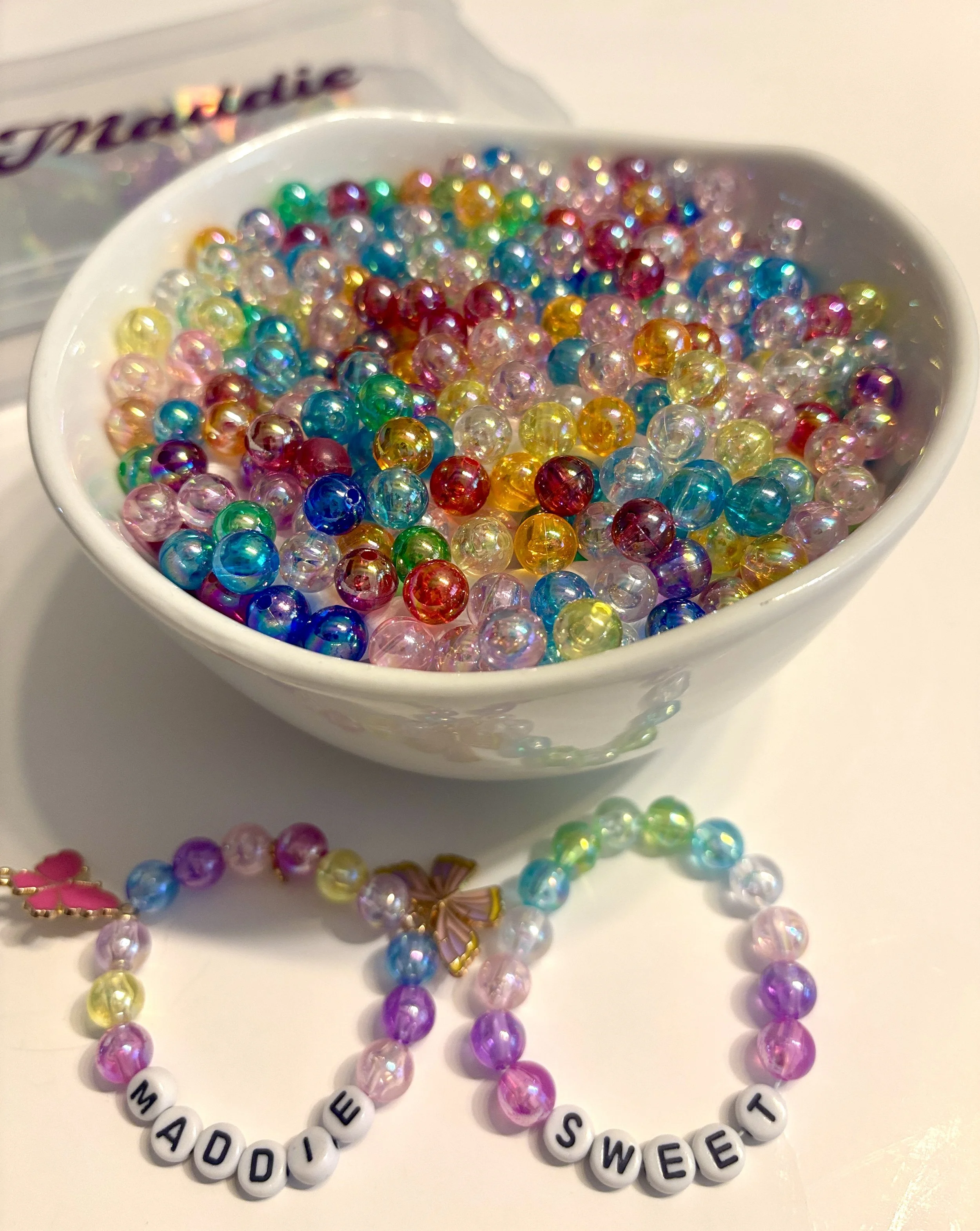 Colorful beaded jewelry, including a bracelet with beads spelling out 'MADDIE'S SWEET' and two rainbow-colored beaded necklaces arranged on a white surface.