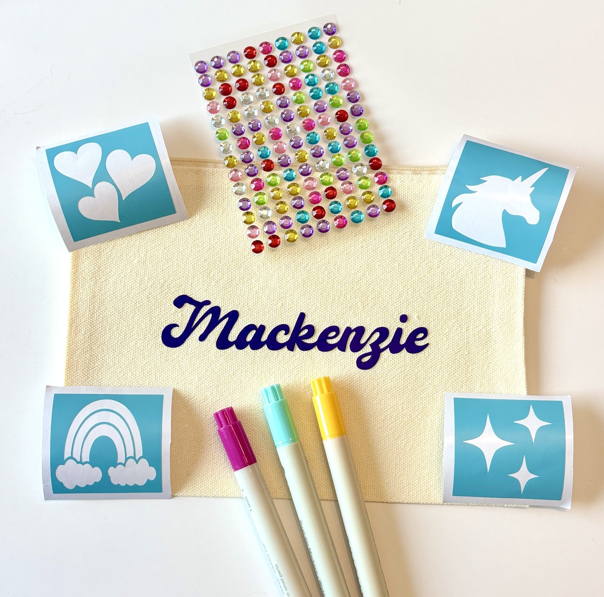 Colorful rhinestones, blue stickers with white hearts and unicorn, and pastel-colored markers are arranged on a beige and white background, spelling out "Thank you".