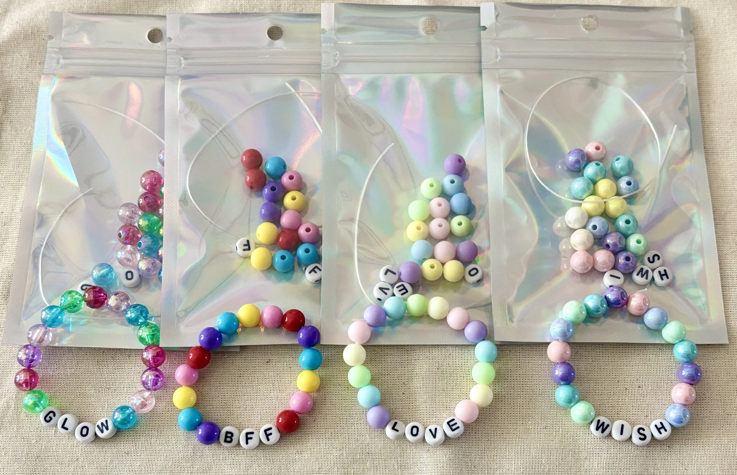 Four plastic bags filled with colorful beads, each with a white string, laid out on a light fabric surface.