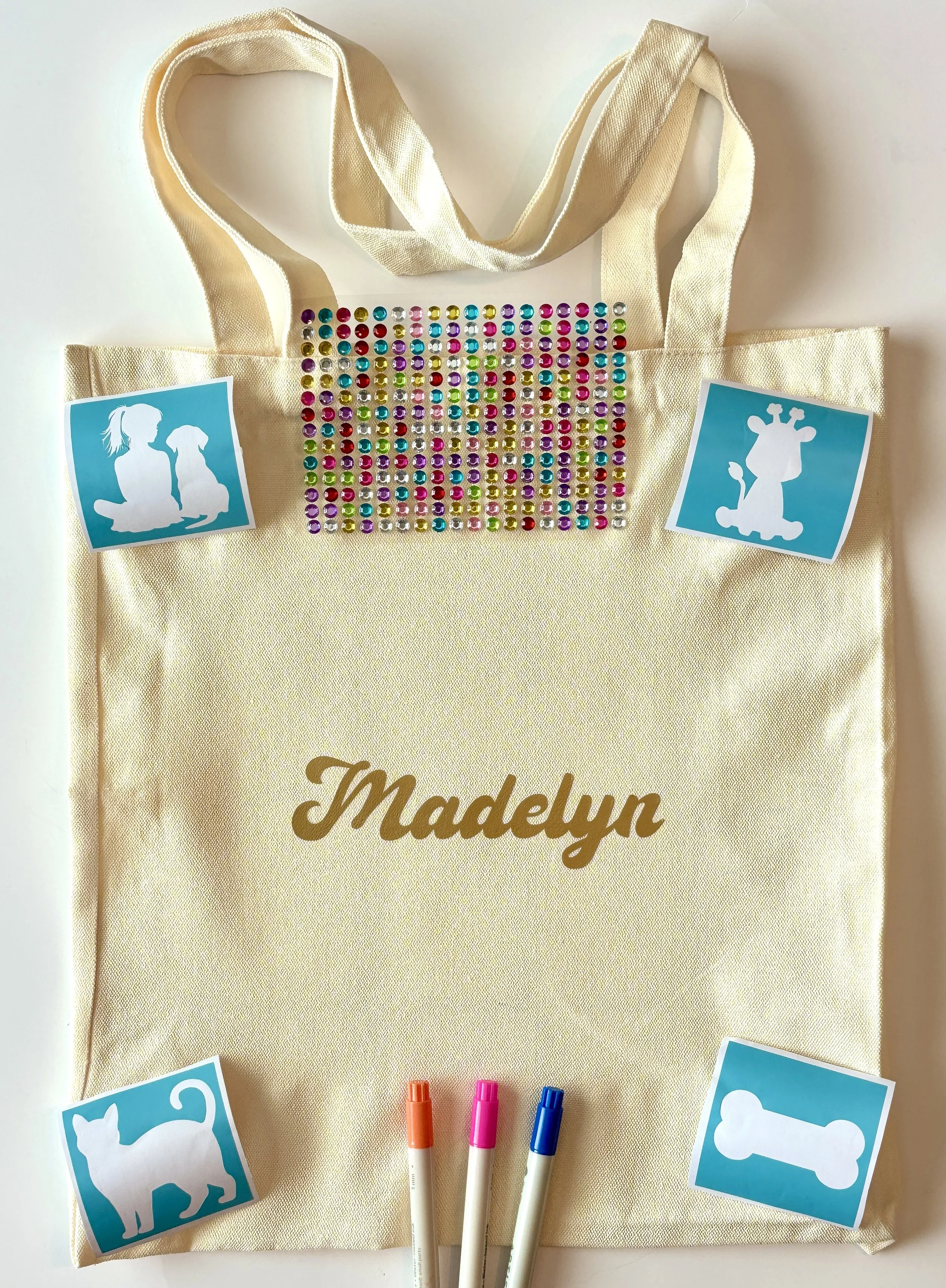 A beige tote bag with the name 'Madelyn' embroidered in gold on it. The tote is decorated with colorful rhinestones in the center and has four dog silhouette stickers in blue and white on the front, along with three markers in orange, pink, and blue 