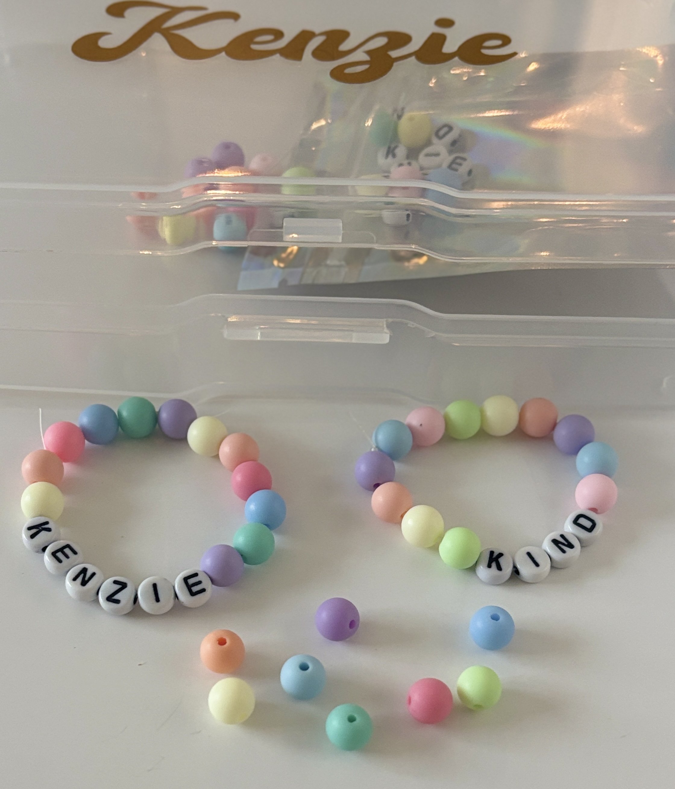Colorful beaded bracelet spelling 'KINDNESS' with pastel and black letter beads, placed on a white surface with scattered pastel beads, and a clear plastic container with a gold script word 'Kenzie' on it.