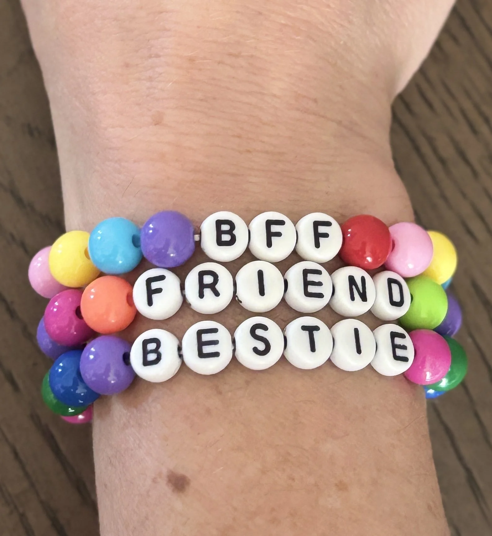 Close-up of multiple colorful bead bracelets on a wrist, with white letter beads spelling ”BFF FRIEND BESIDE”