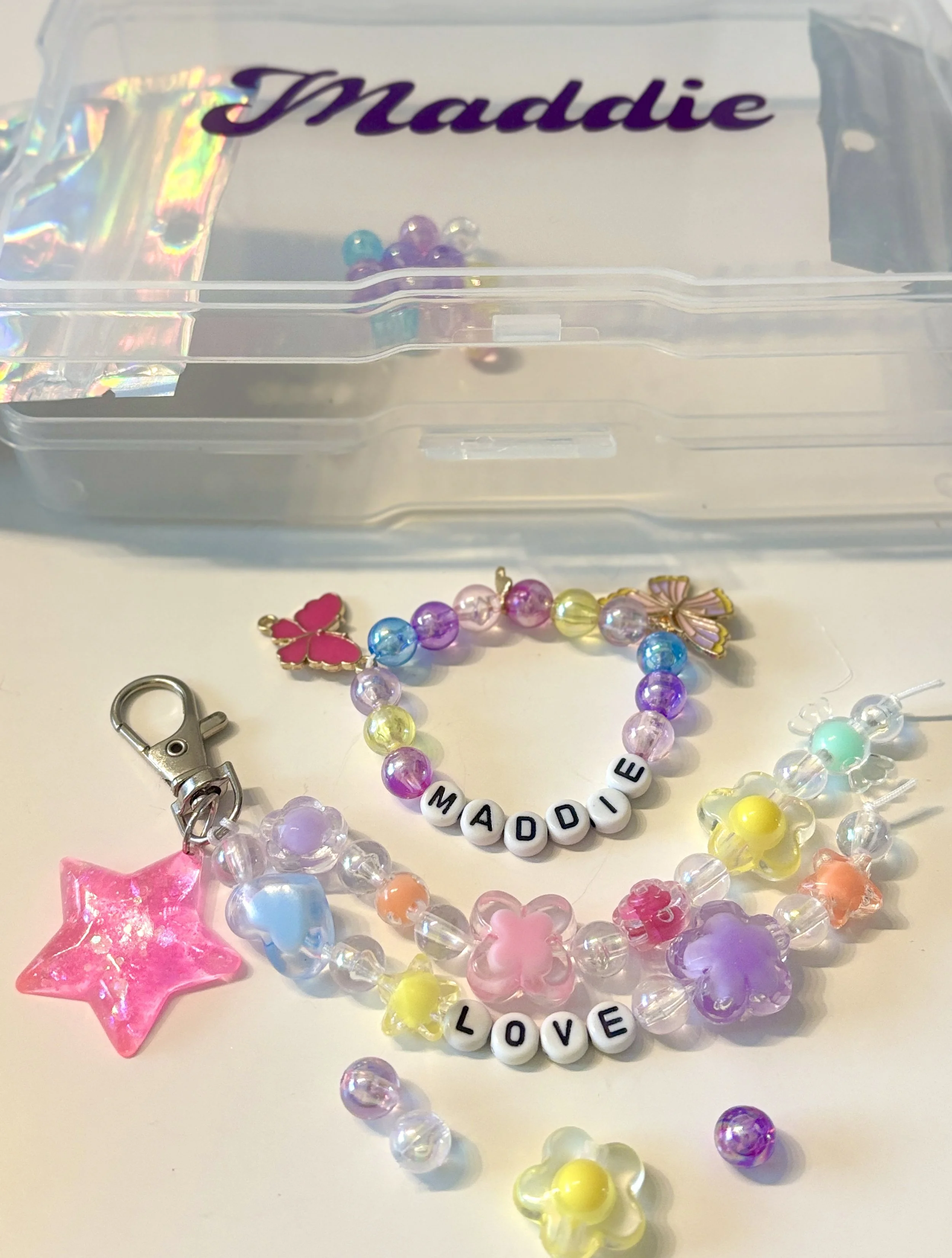 Colorful beaded jewelry with pearl-like beads and word beads spelling out "MADDI" and "LOVE," along with star and butterfly attachments, and a plastic container in the background labeled "Maddi."