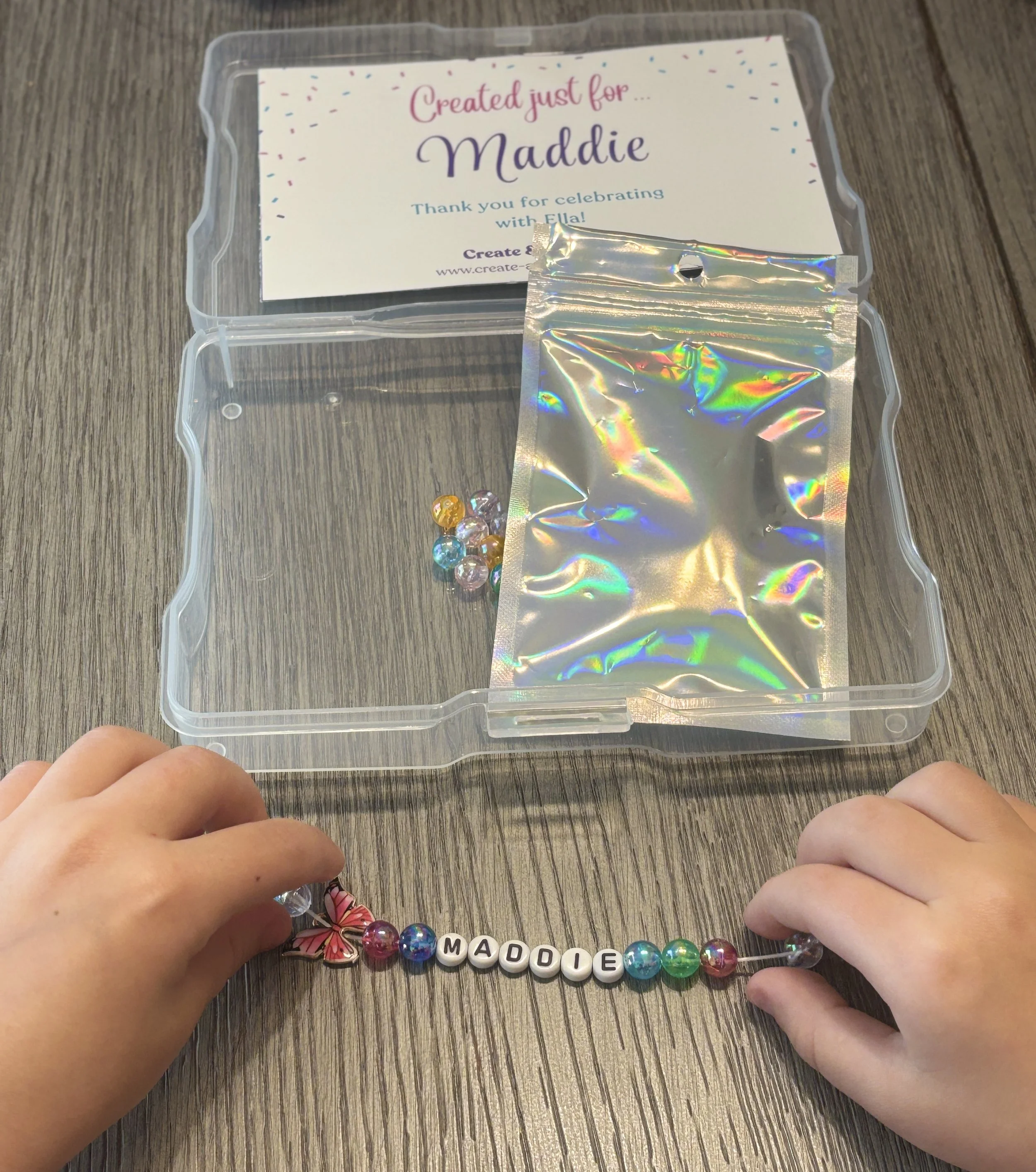 Child's hands holding a beaded bracelet spelling 'MADDIE' inside a plastic box on a wooden surface.