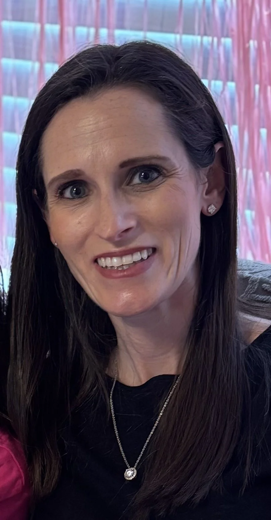 A woman with long dark hair, blue eyes, and a friendly smile, wearing a black top, diamond earrings, and a necklace, in front of a colorful, neon-lit background.