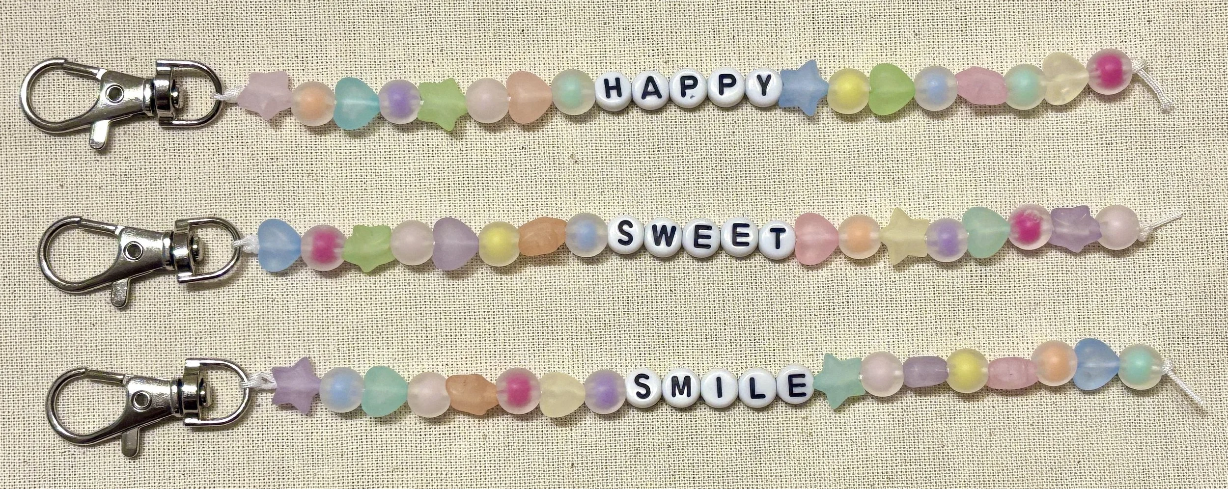 Colorful beaded keychains with pastel stars, hearts, and beads, spelling out 'HAPPY,' 'SWEET,' and 'SMILE' on a beige fabric background.