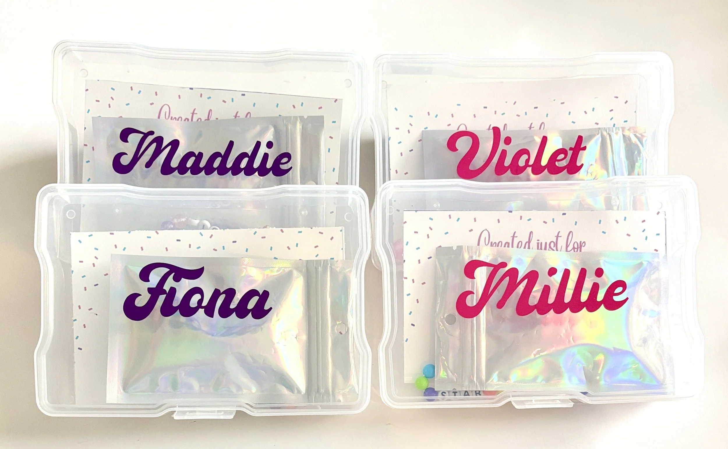 Clear plastic storage containers holding personalized labels for various names, such as Maddie, Violet, Fiona, Millie, and others, with decorative confetti-like designs and holographic accents.