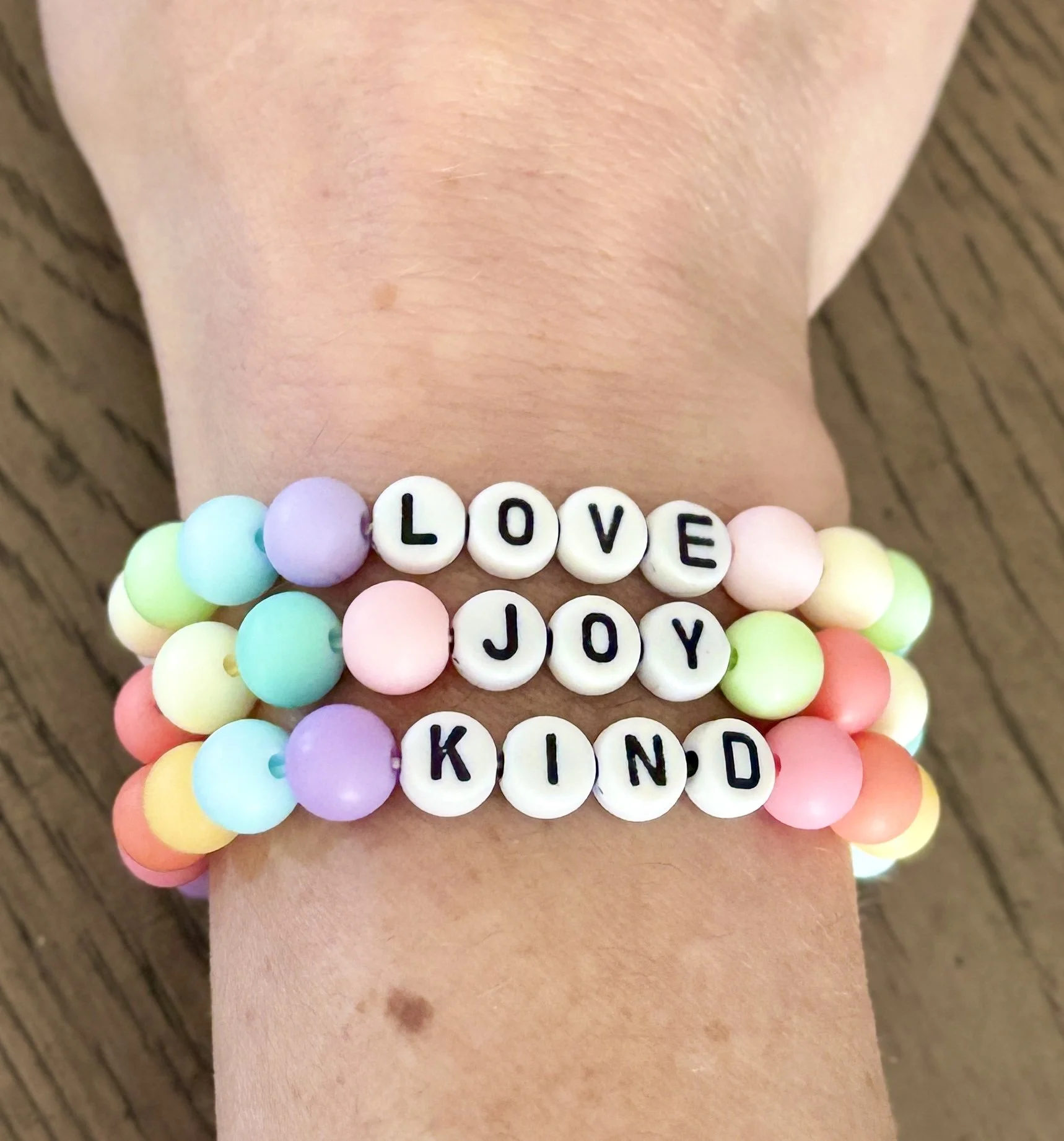 Colorful beaded bracelet with white beads spelling out 'LOVE,' 'JOY,' and 'KIND,' worn on a person's wrist over textured wooden background.