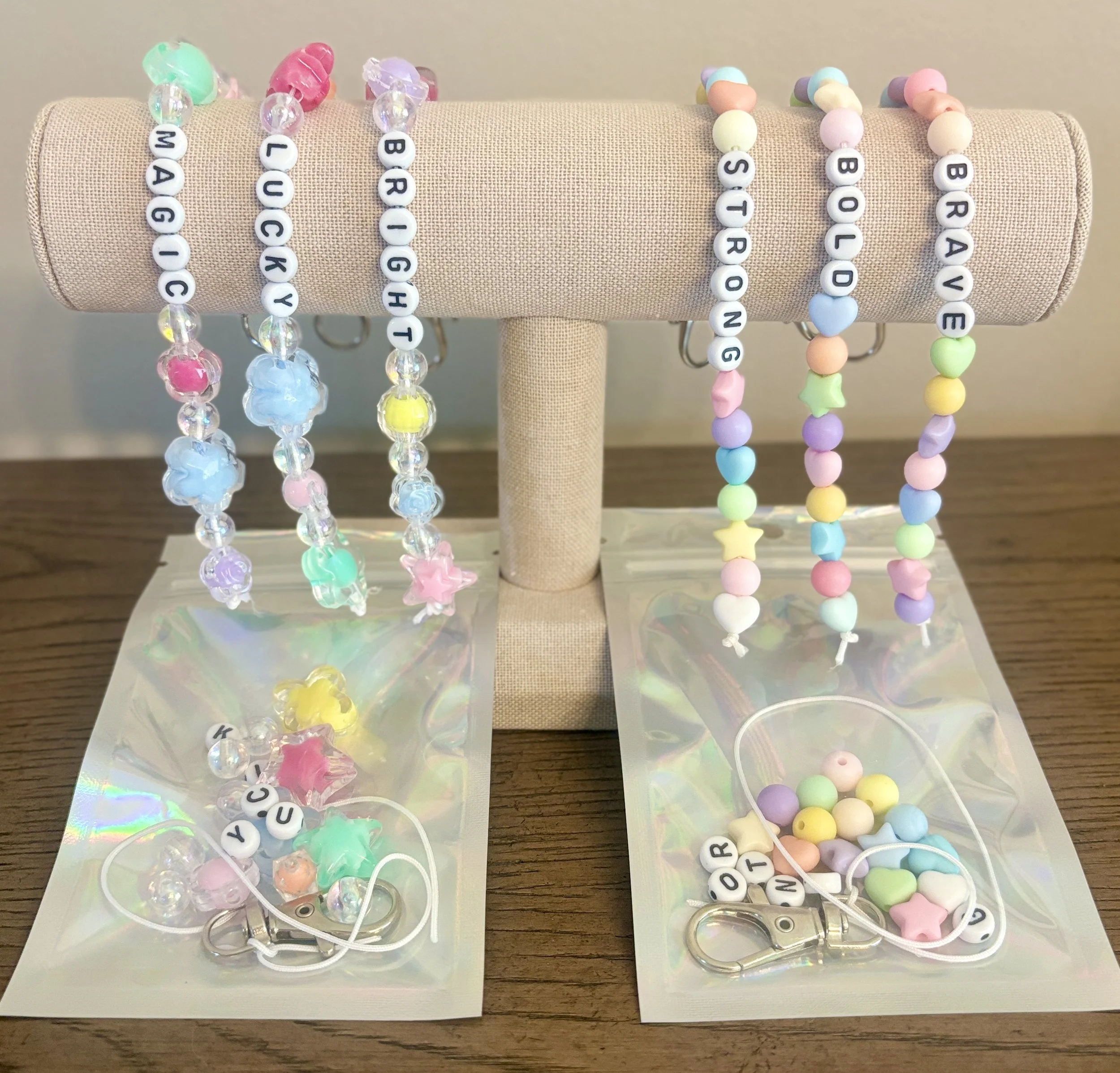 Colorful beaded bracelets with letter beads spelling names or words, displayed on a beige fabric-covered stand, with some beads shaped like hearts, stars, and animal shapes, on a wooden surface.