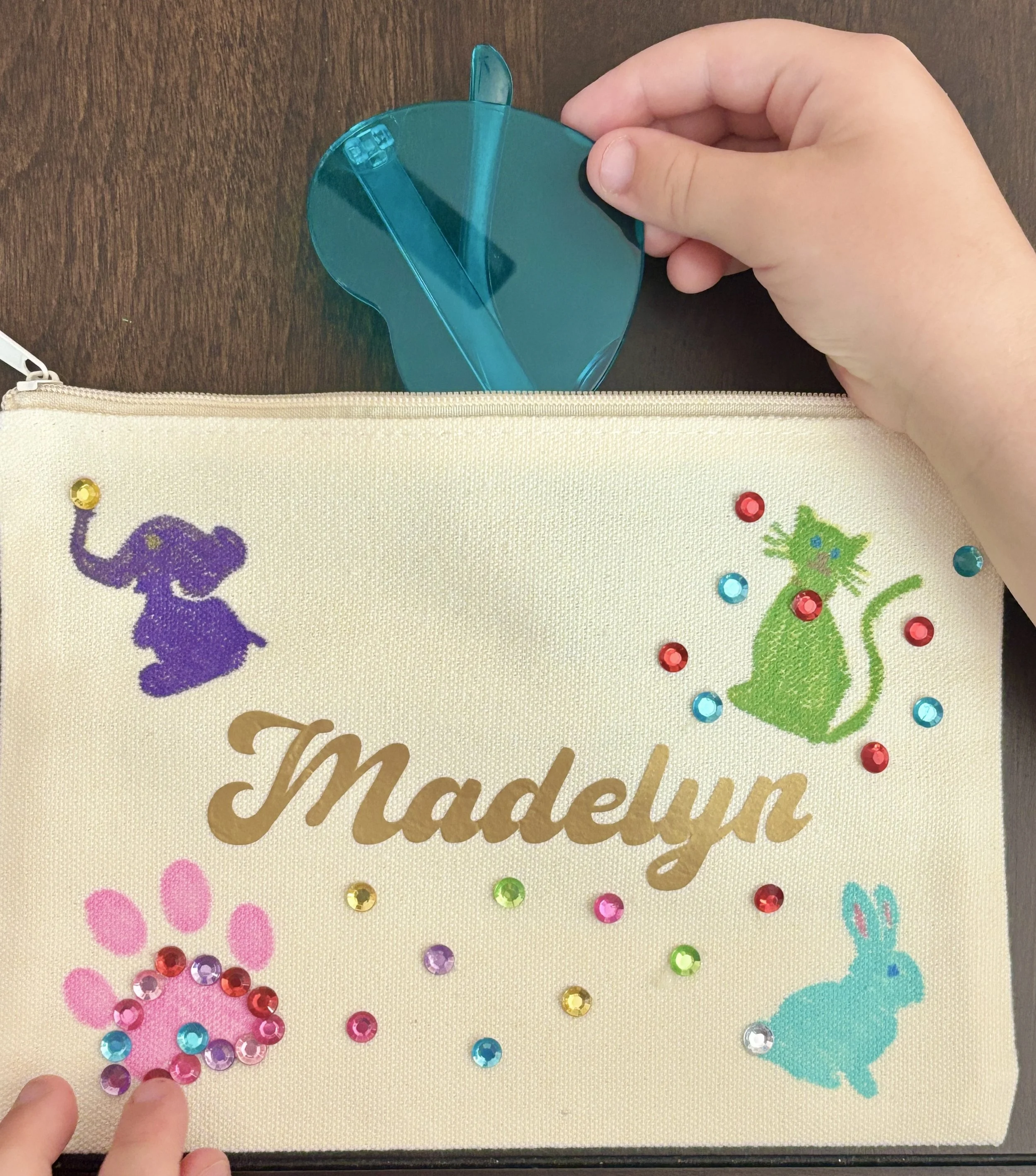 A hand placing a teal plastic piece into a white fabric with colorful embroidered animal designs and scattered rhinestones. The fabric has the words 'The Madelina' embroidered in gold.