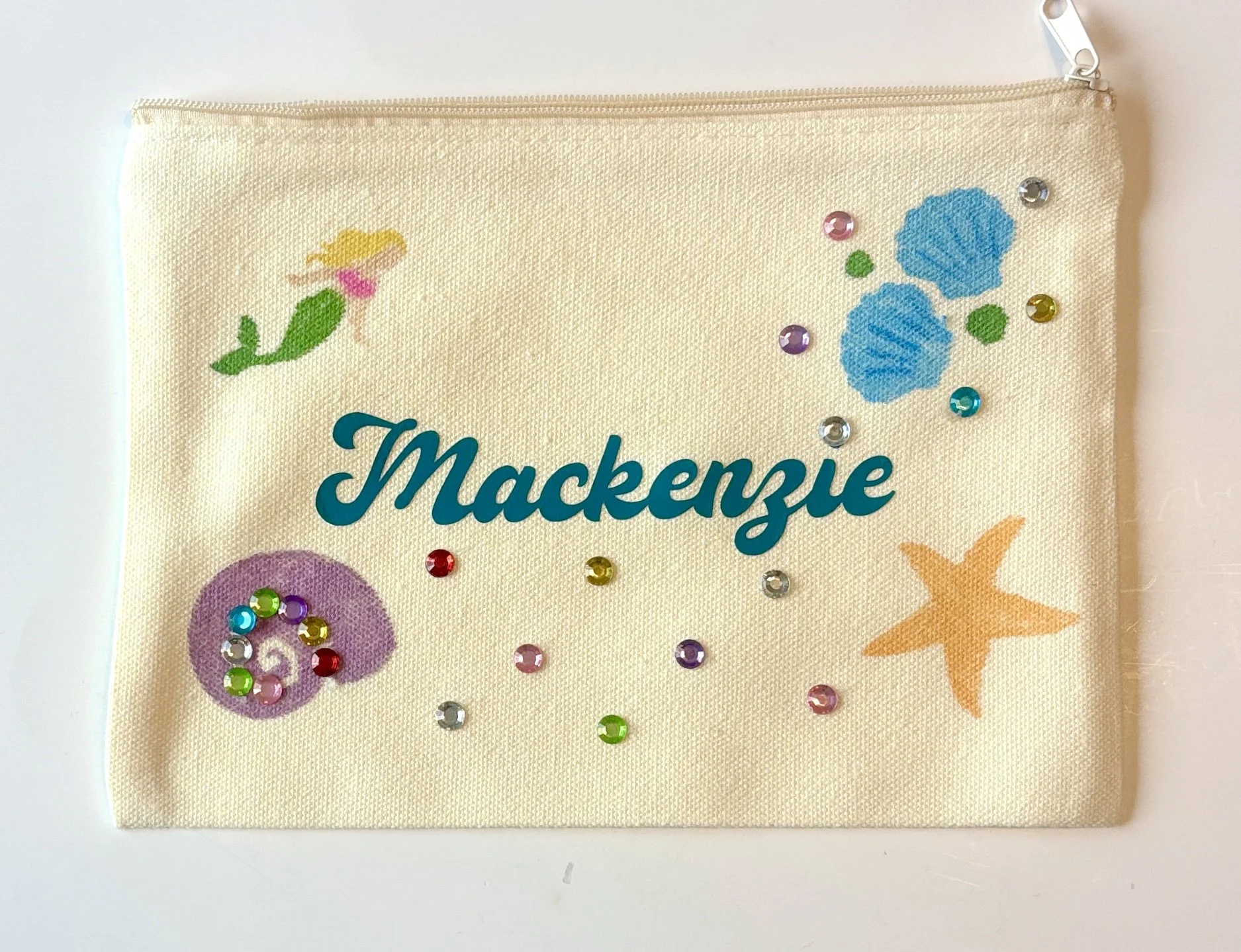 A cream-colored canvas pouch with the name 'Mackenzie' in teal cursive letters, decorated with a small yellow-haired fairy, blue seashells, an orange starfish, colorful rhinestones, and glitter designs.