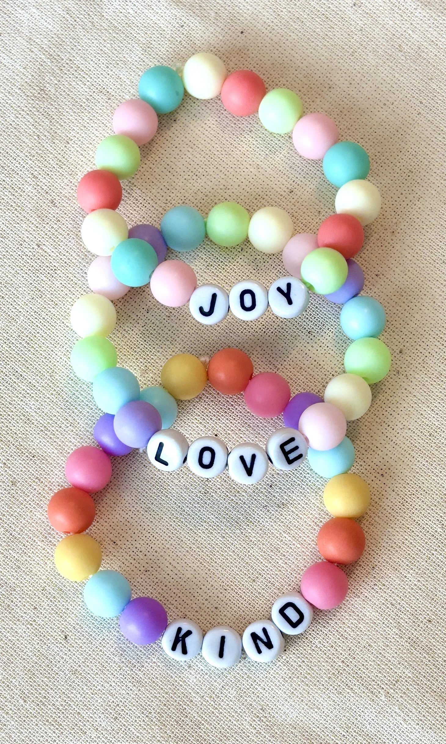 Colorful bead bracelet with letter beads spelling out words like 'JOY', 'LOVE', 'KIND', on a textured beige surface.