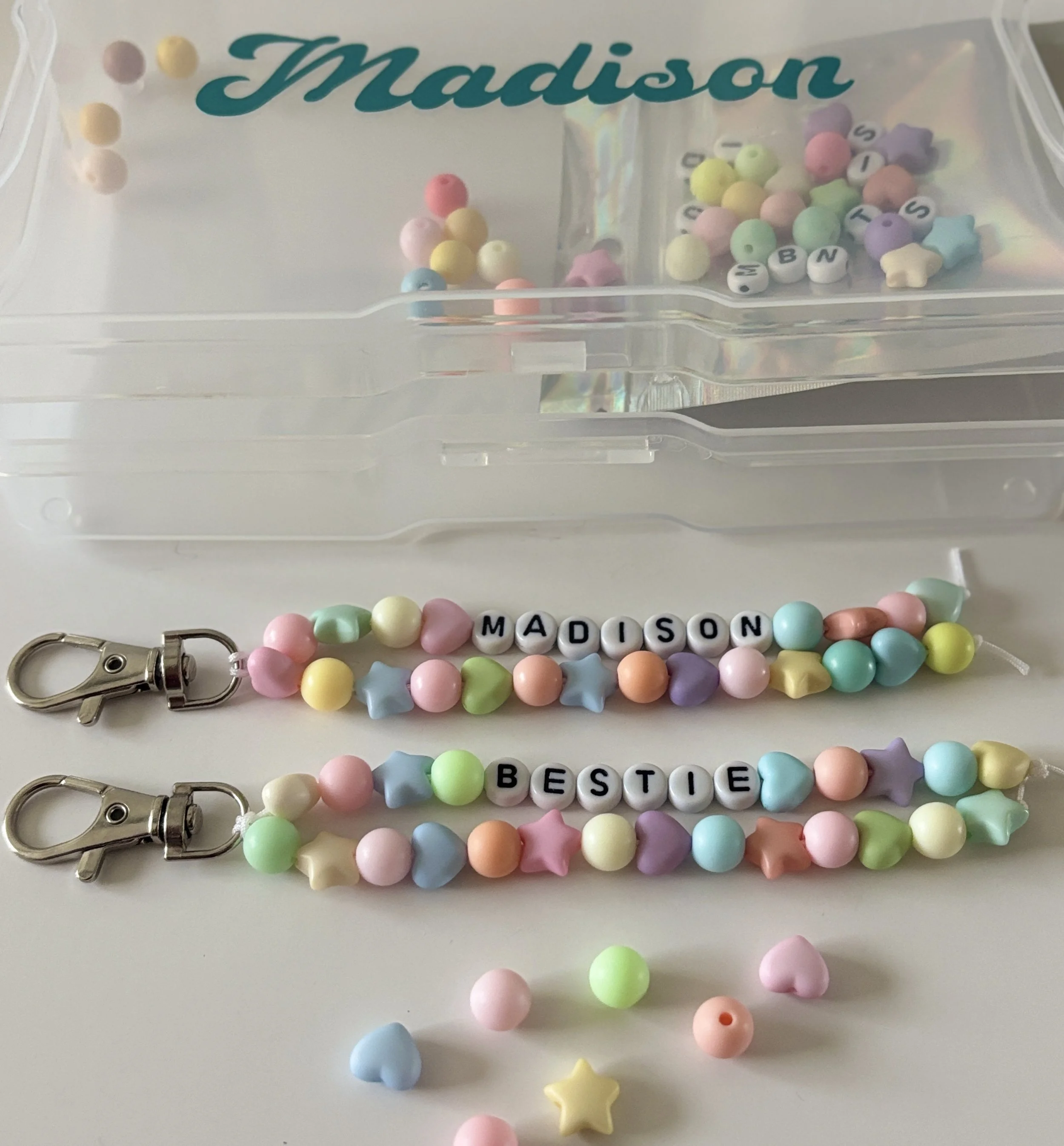 Colorful beaded keychains with letter beads spelling 'Madison' and 'Bestie', with a clear storage box labeled 'Madison' in the background.