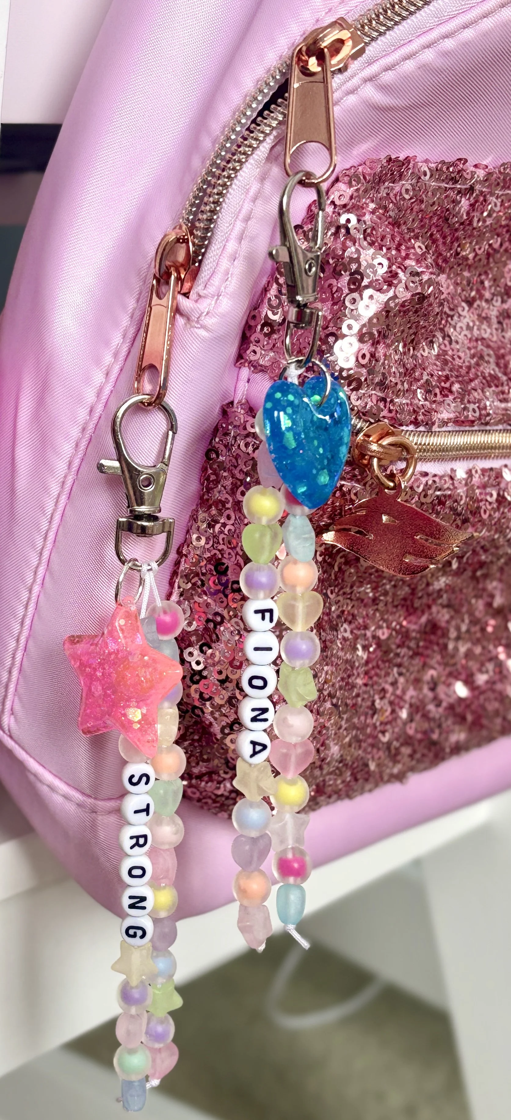 Close-up of pink sequin and satin backpack with decorative beaded accessories hanging from zipper clips.