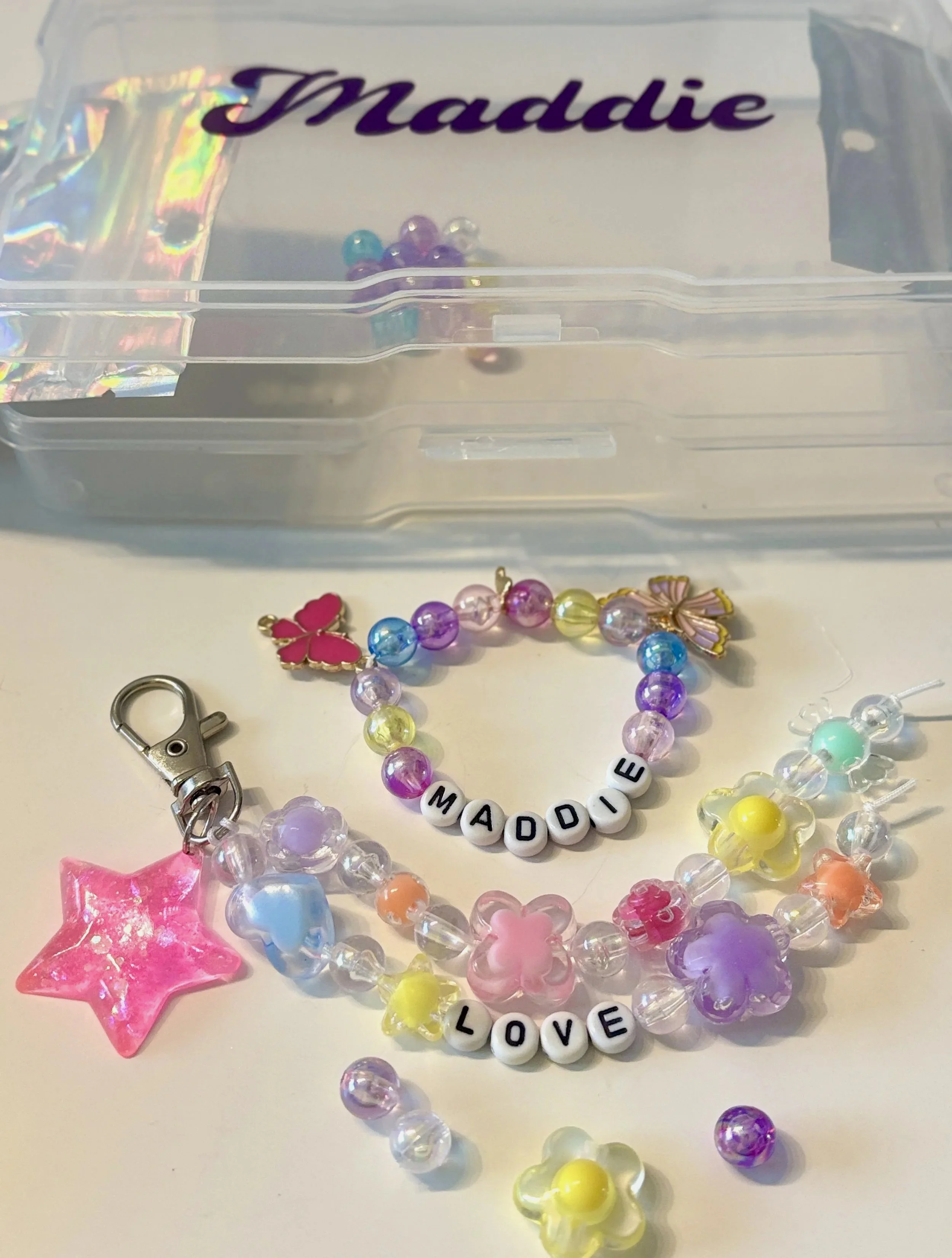 Colorful beaded jewelry pieces with words 'MADDI' and 'LOVE' on beads, pink star-shaped item, detached jewelry parts, and a small metal clip, all laid out on a white surface.